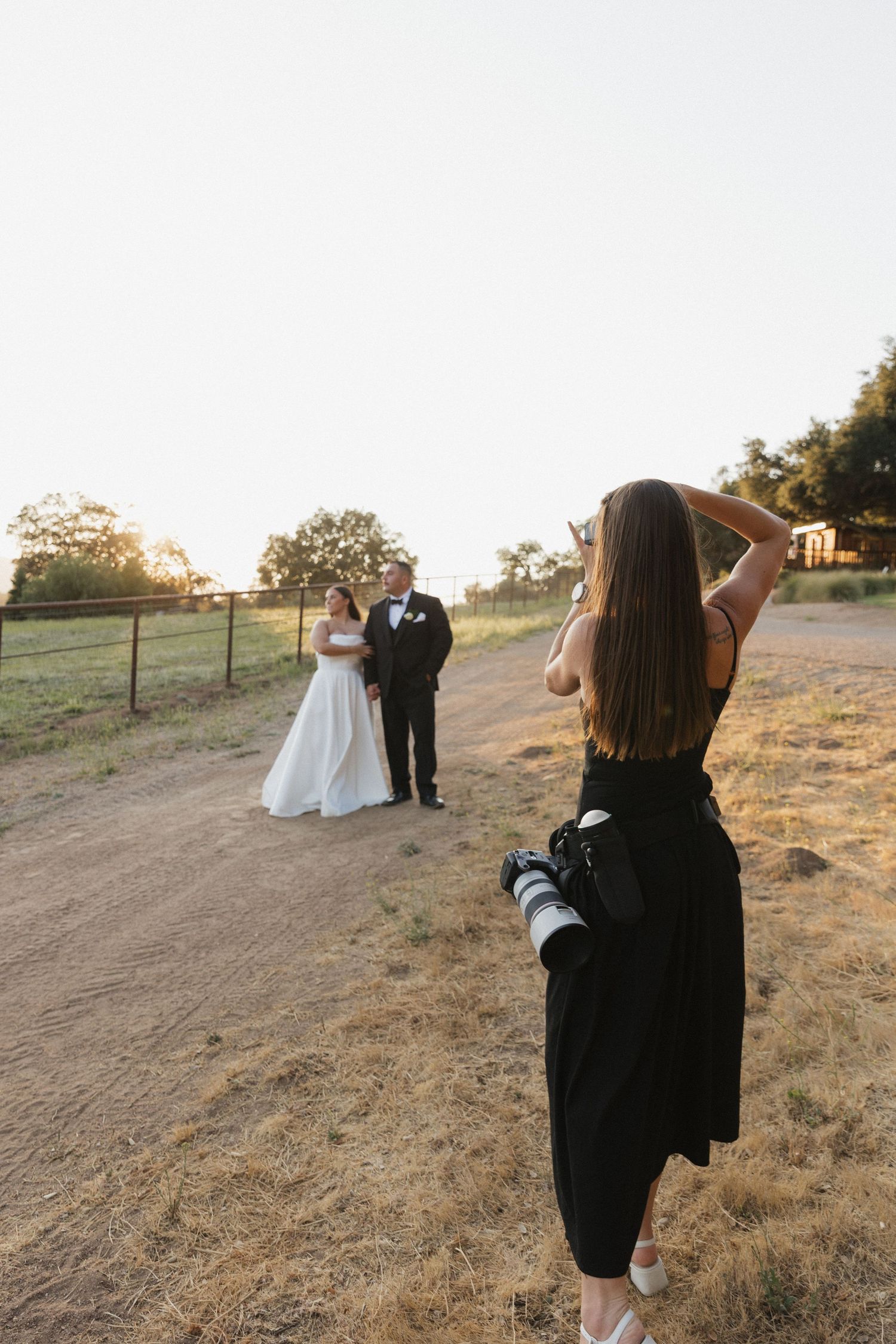 Wedding photographer captures couple on a dirt road during golden hour sunset at rustic outdoor venue.