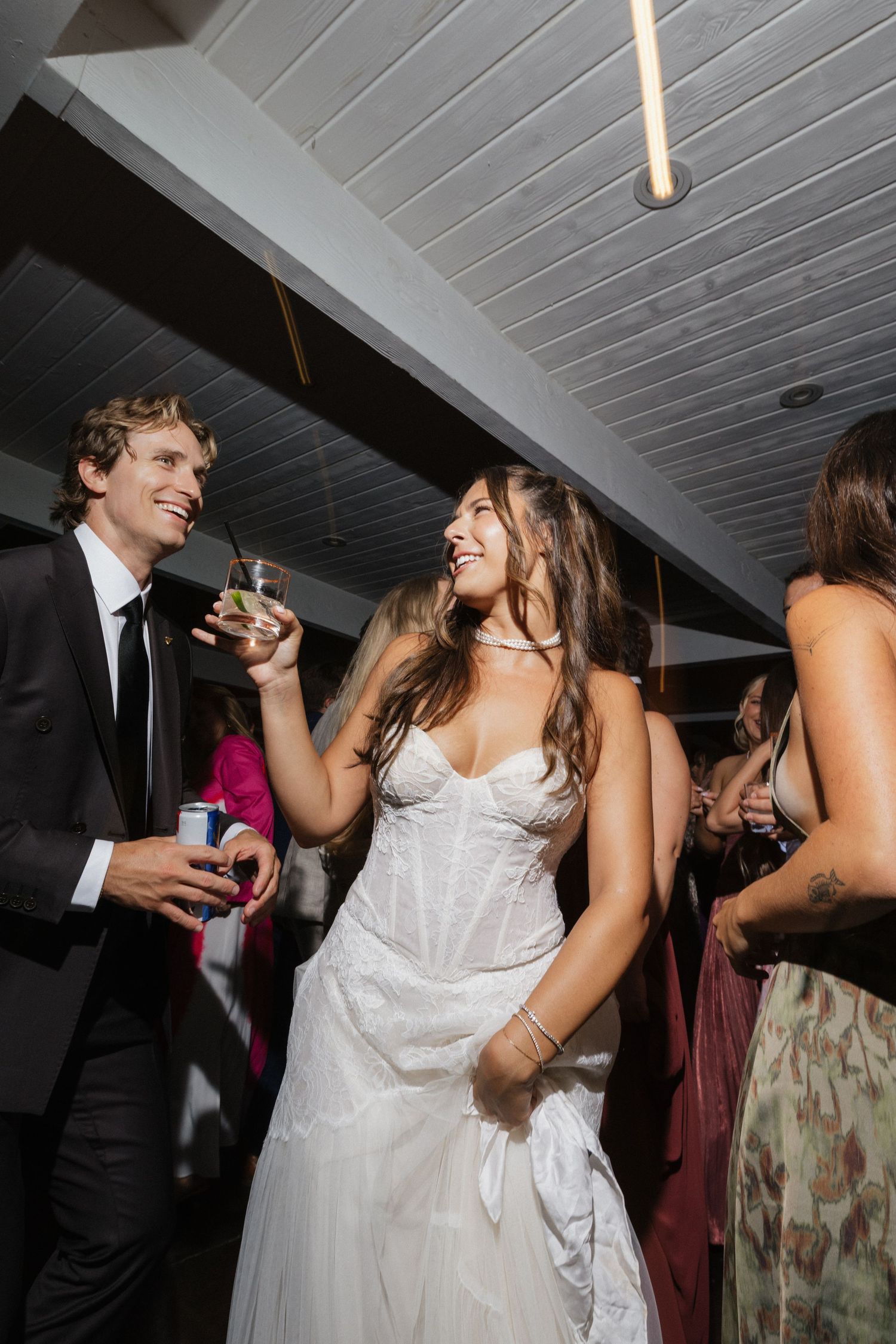 Bride in white gown laughing with groom holding drinks at wedding reception.