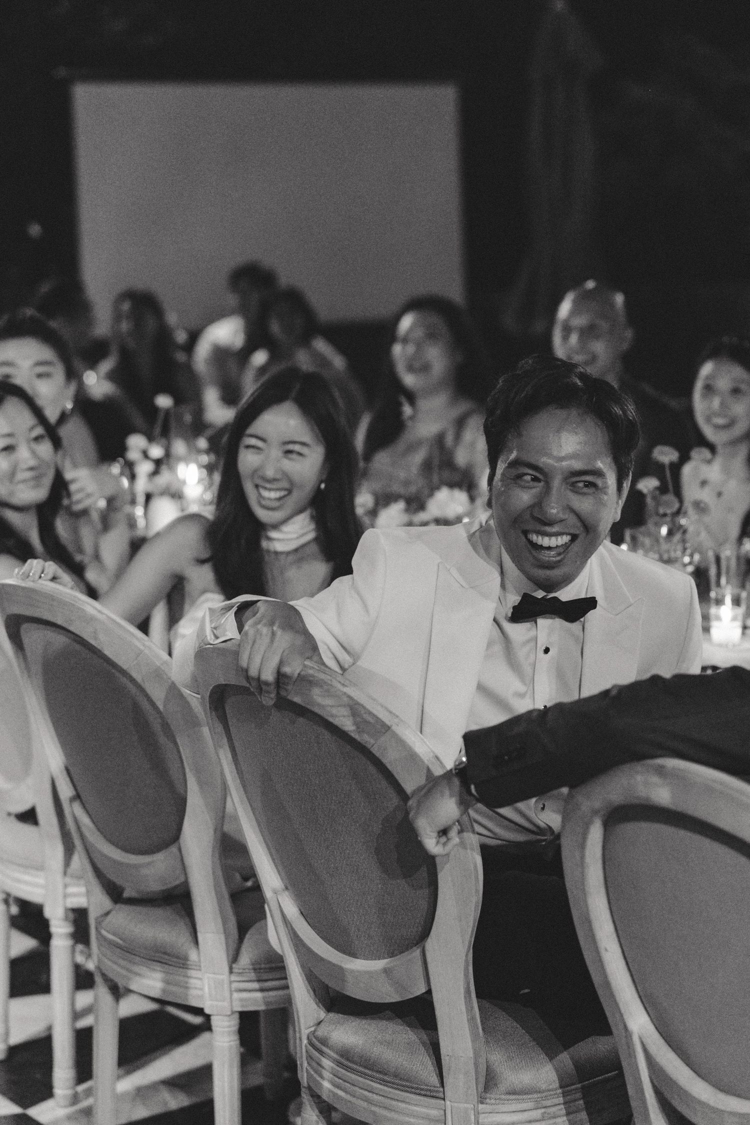 Wedding guests seated in elegant chairs laughing during reception celebration.