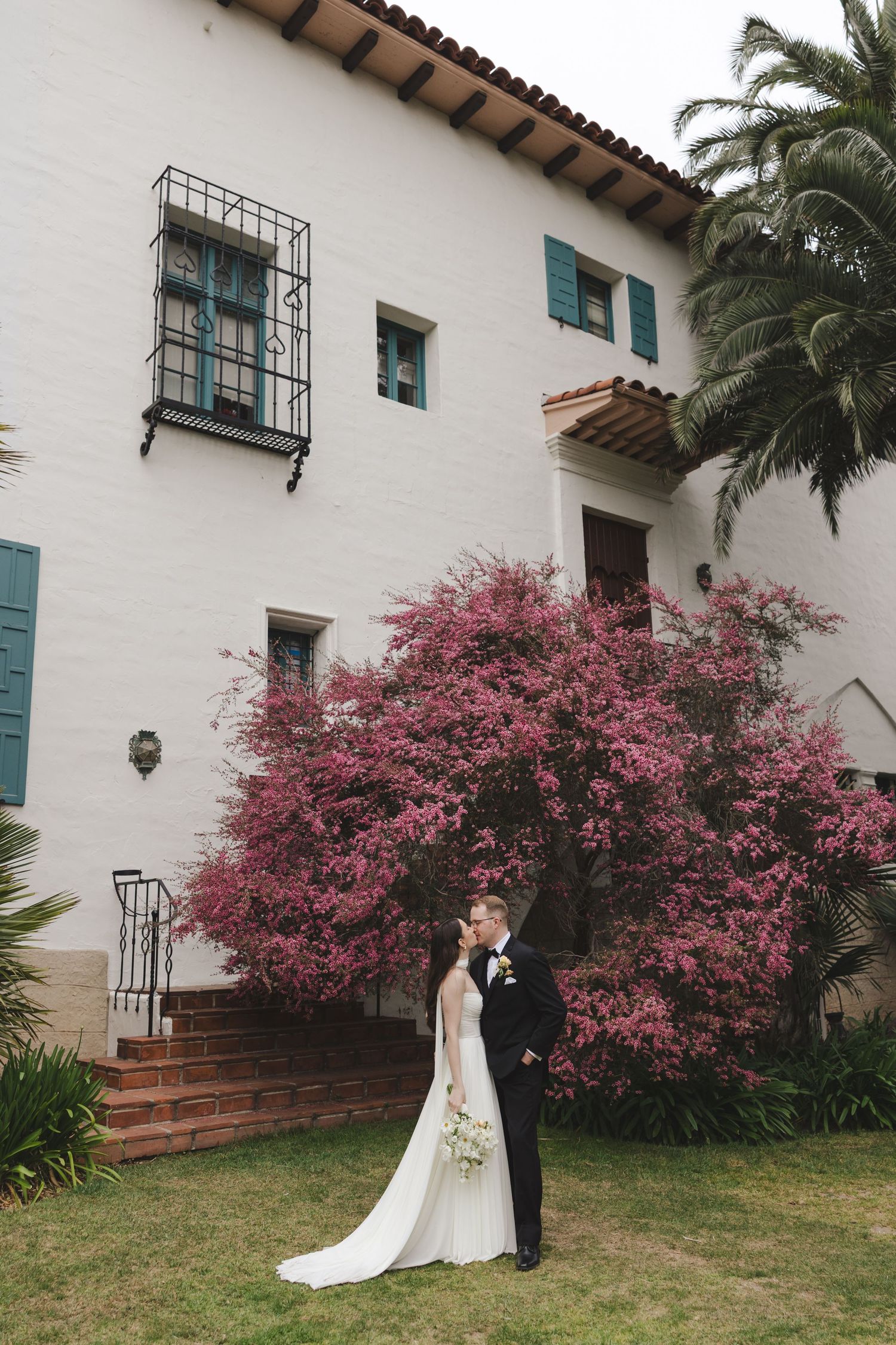 Spanish colonial villa with white walls, blue shutters and blooming pink bougainvillea frames couple in wedding attire.