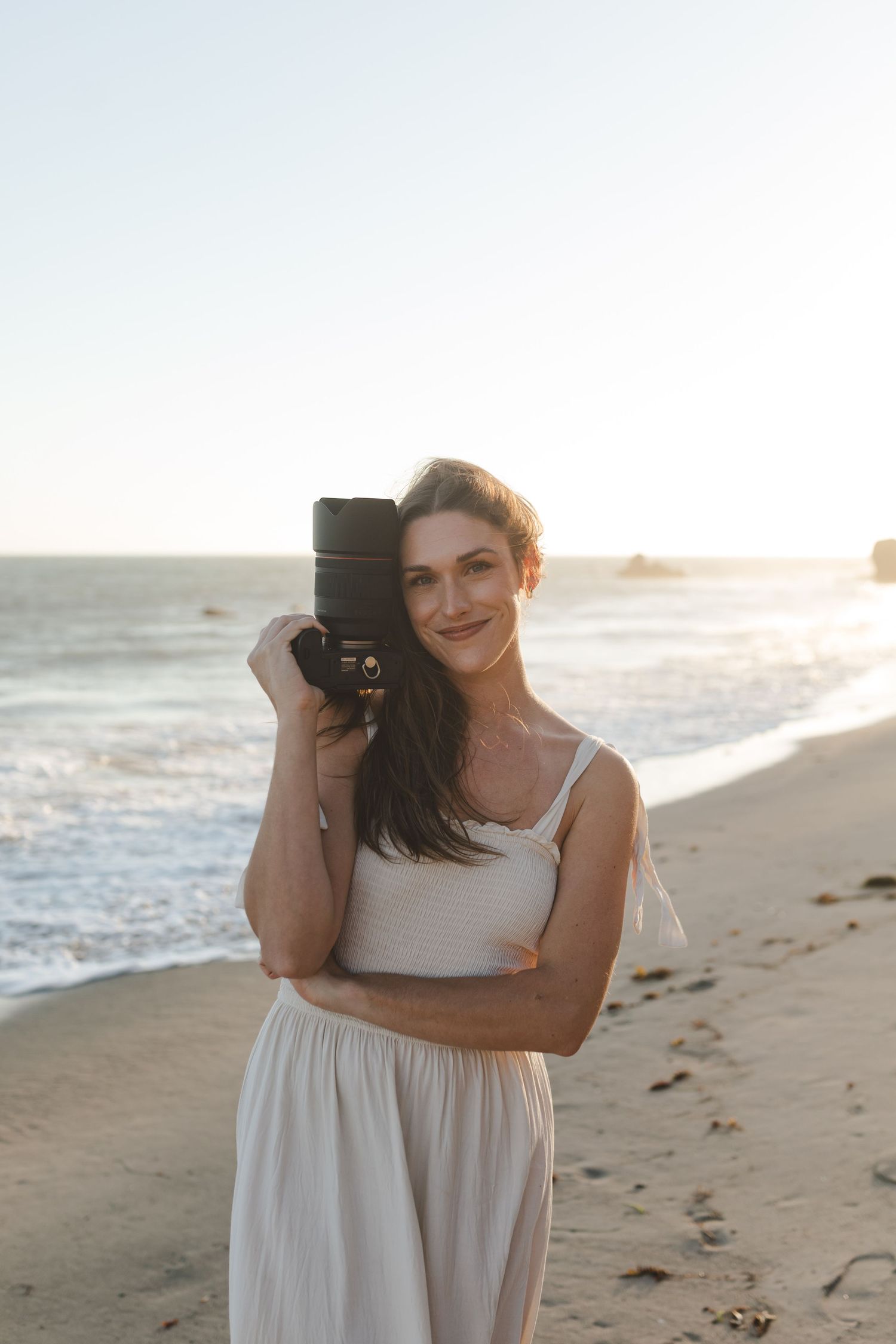 A photographer in a white dress holds a professional camera on a sunlit beach at golden hour.