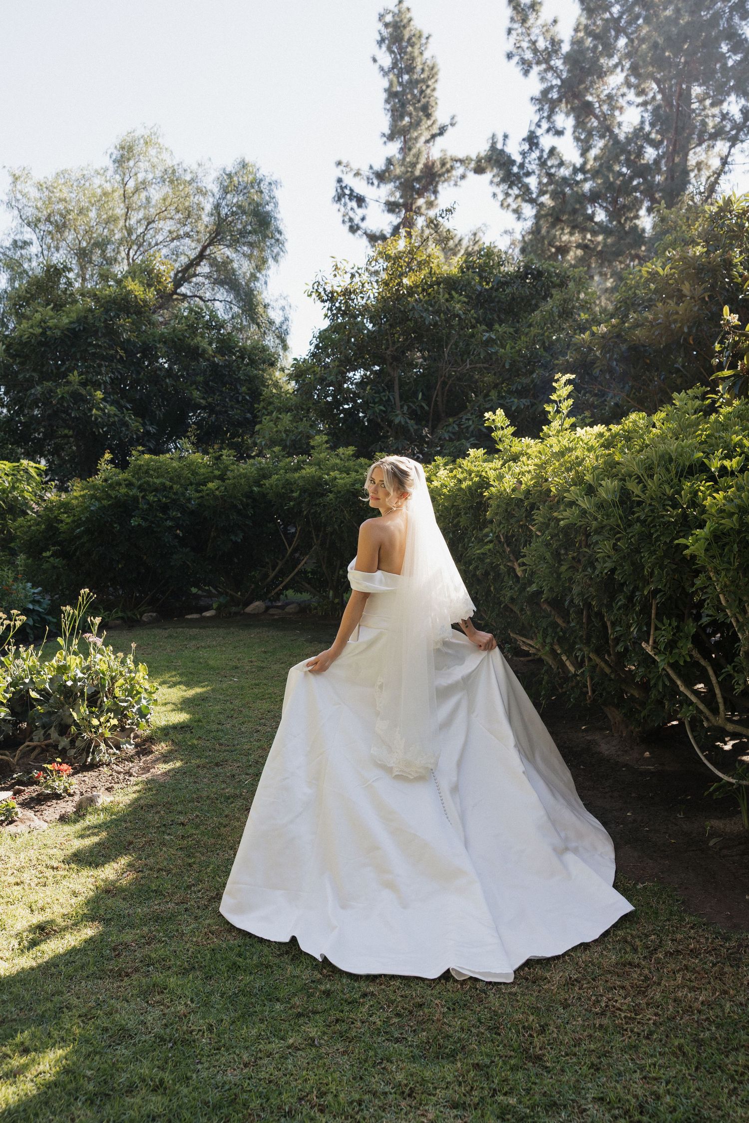 An elegant wedding dress with a flowing train photographed in a sunlit garden setting.