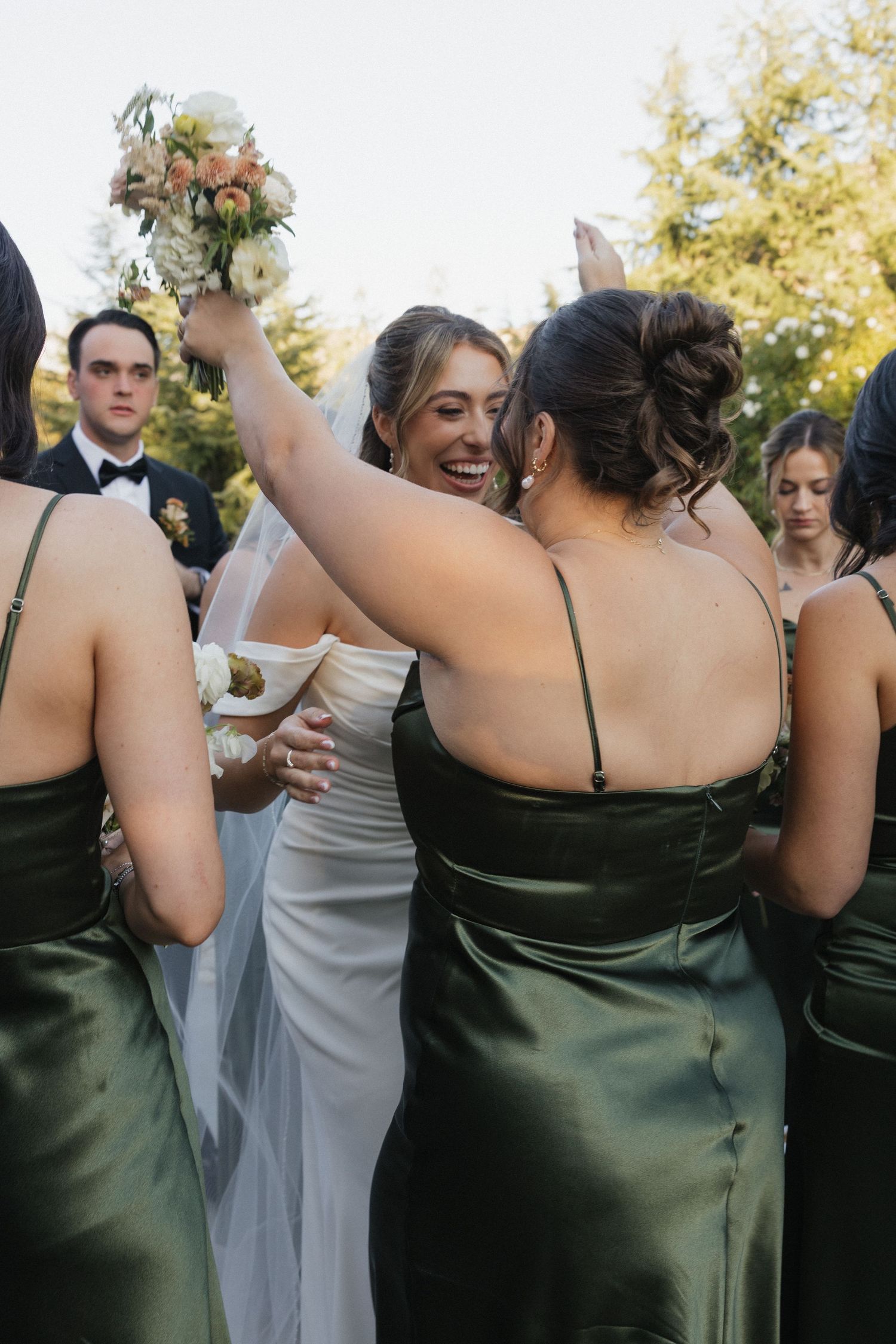 Wedding party members in dark green dresses and white gown celebrate with hugs and laughter outdoors.