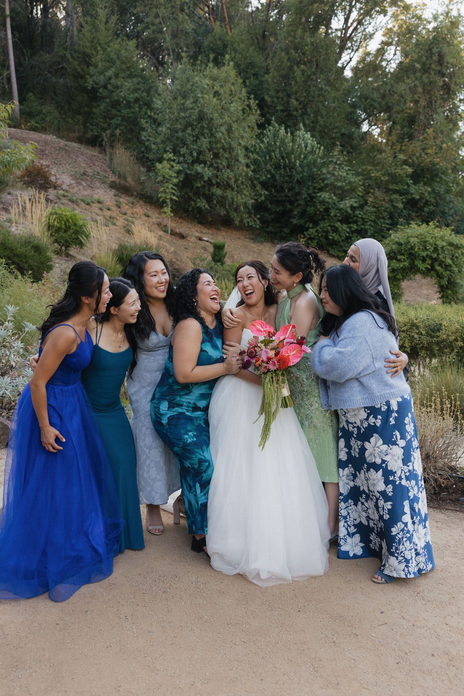 A bridal party in blue and floral dresses poses together outdoors on a sunny day.