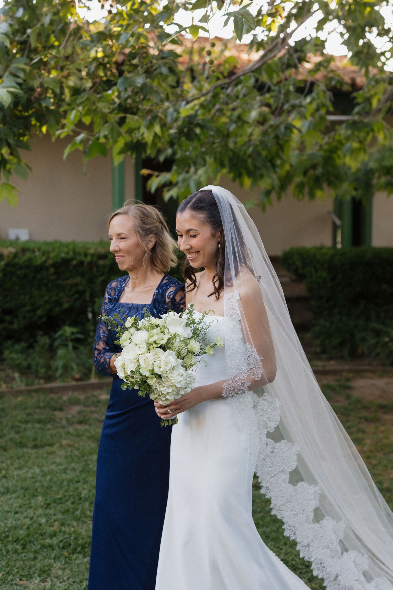 Bride in white gown and veil holds white bouquet beside older woman in navy lace dress outdoors.