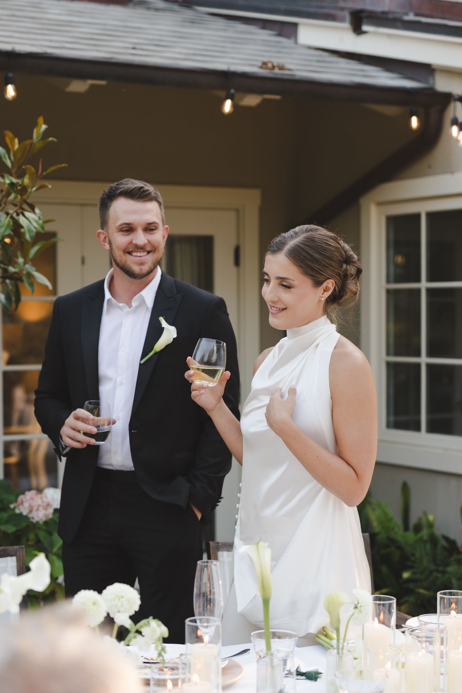 Wedding guests enjoy drinks and conversation at an elegant outdoor reception with string lights overhead.