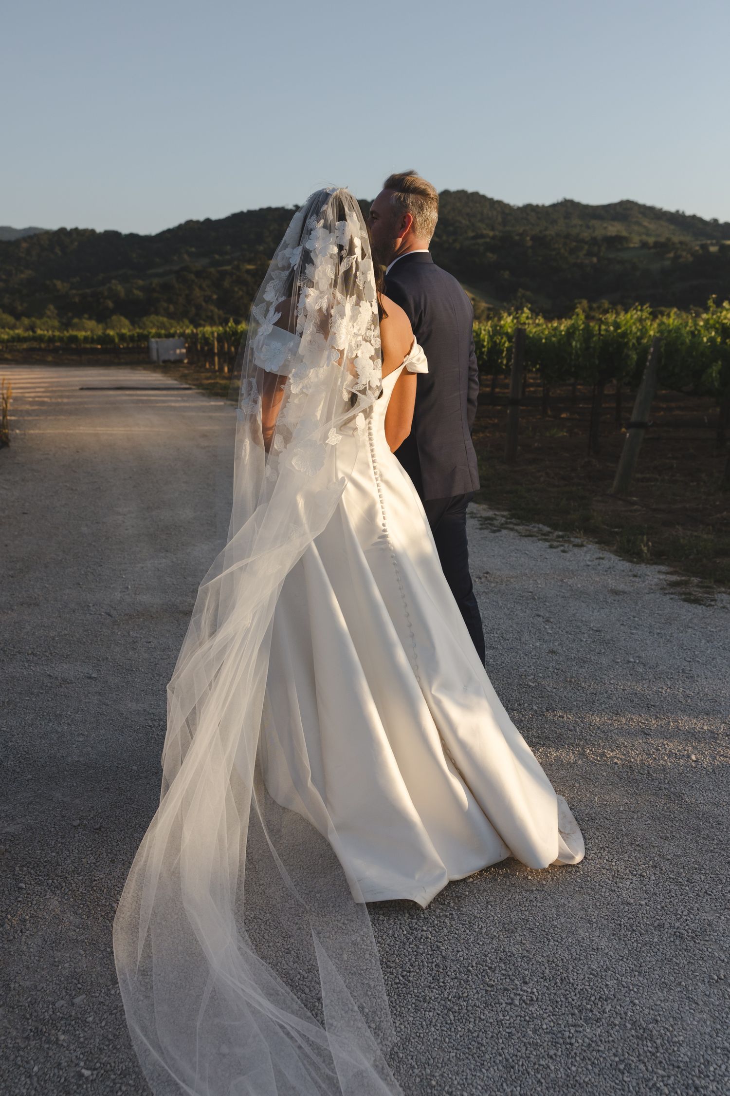 Newlyweds walk together on gravel path with flowing white dress and veil.