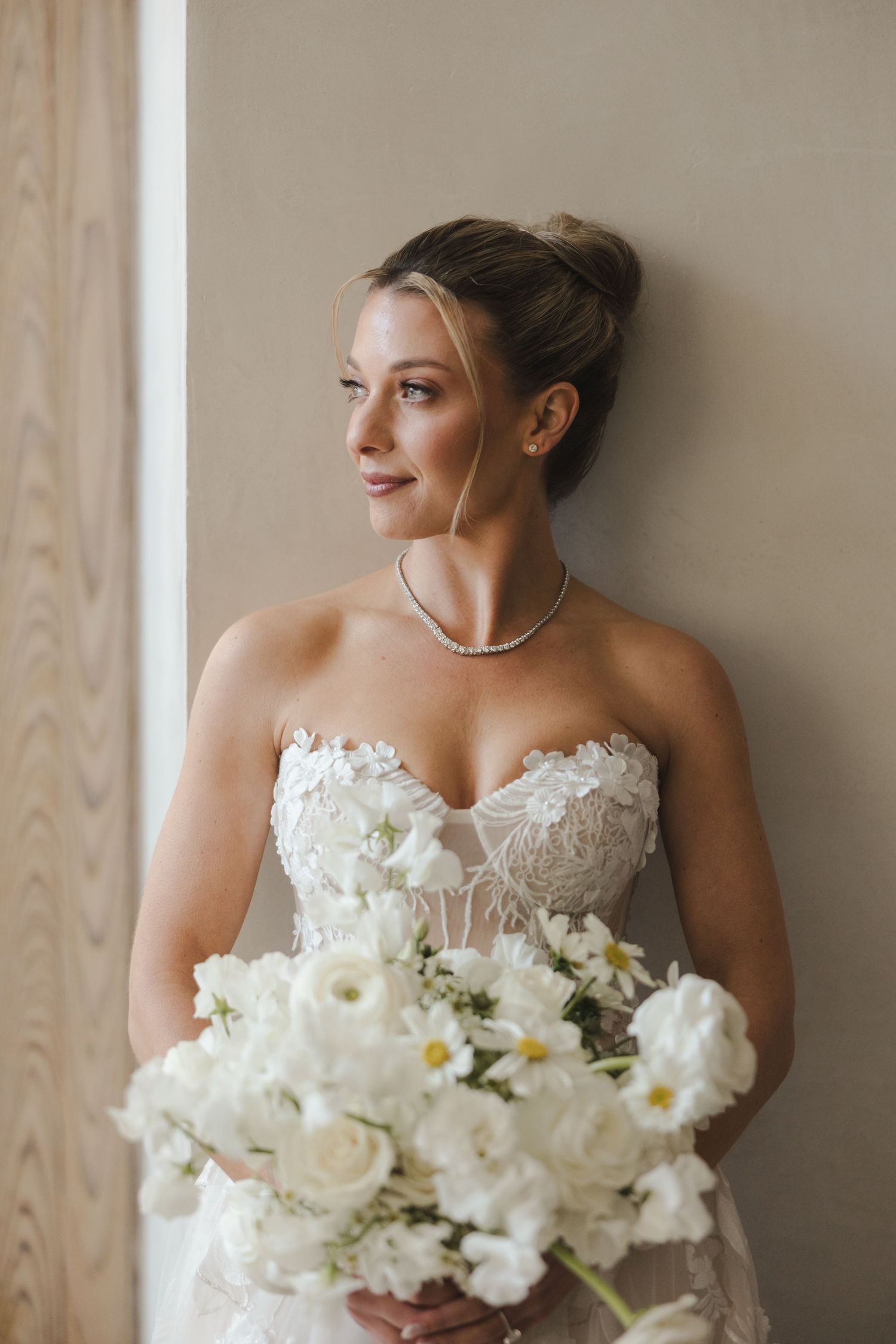 A bride in a strapless white lace gown holds a bouquet of white roses and daisies while gazing out a window.