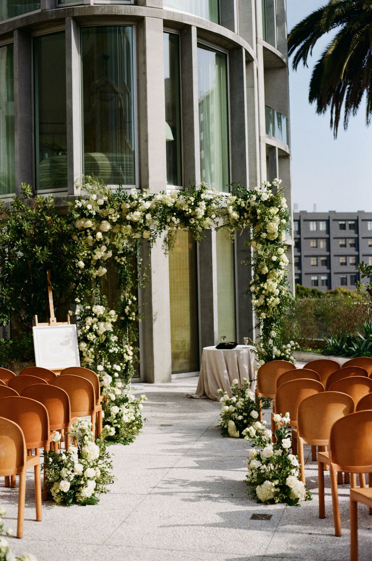 Modern outdoor wedding ceremony setup with brown chairs and white floral arrangements along a glass building entrance.