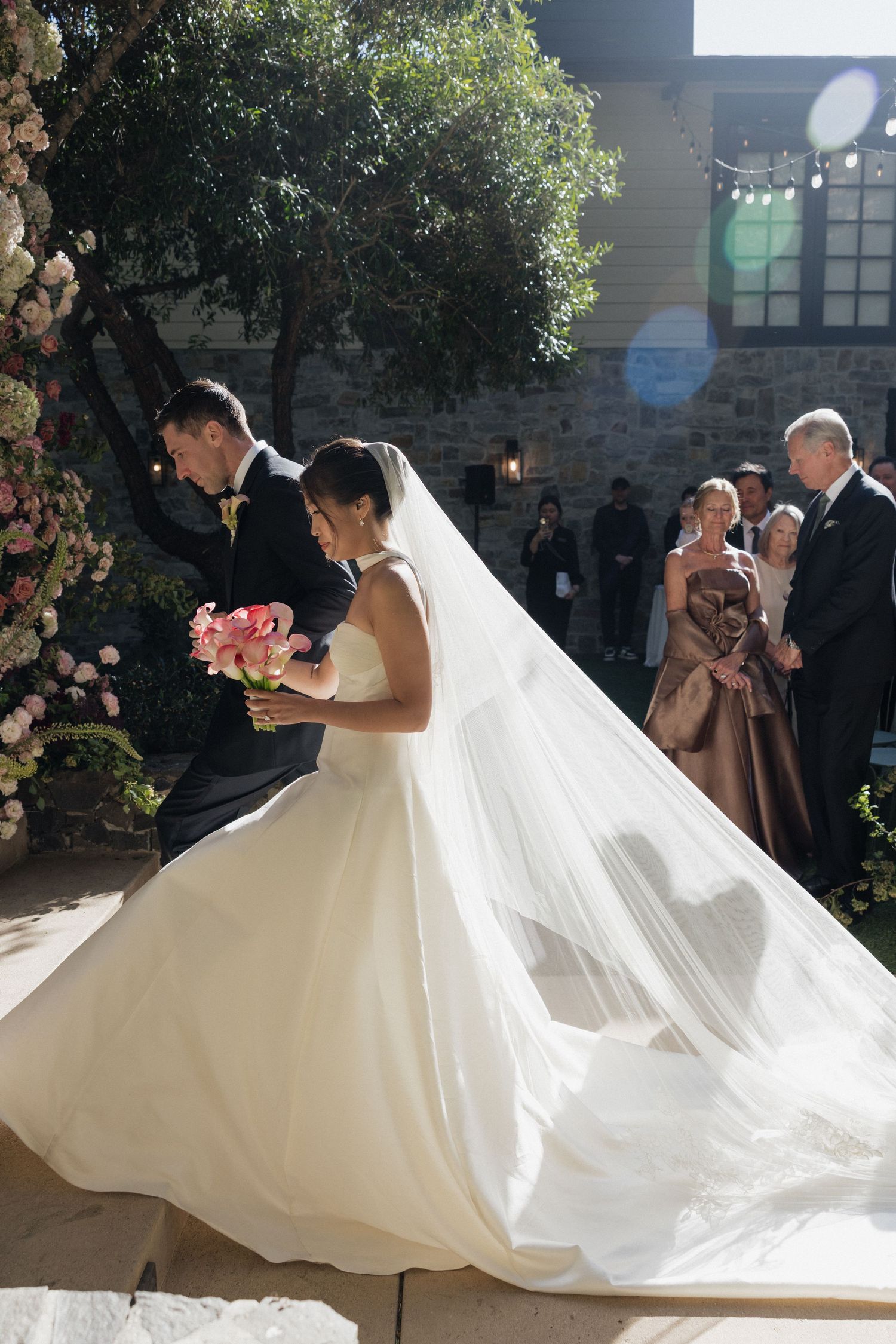 Bride in flowing white gown with long veil walks with groom at outdoor wedding ceremony surrounded by floral arrangements.