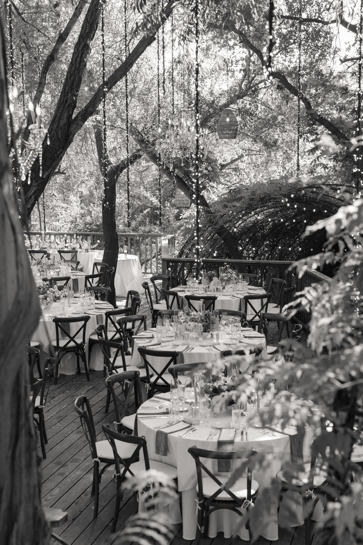 Black and white photo of elegant outdoor wedding reception with round tables under trees with string lights.