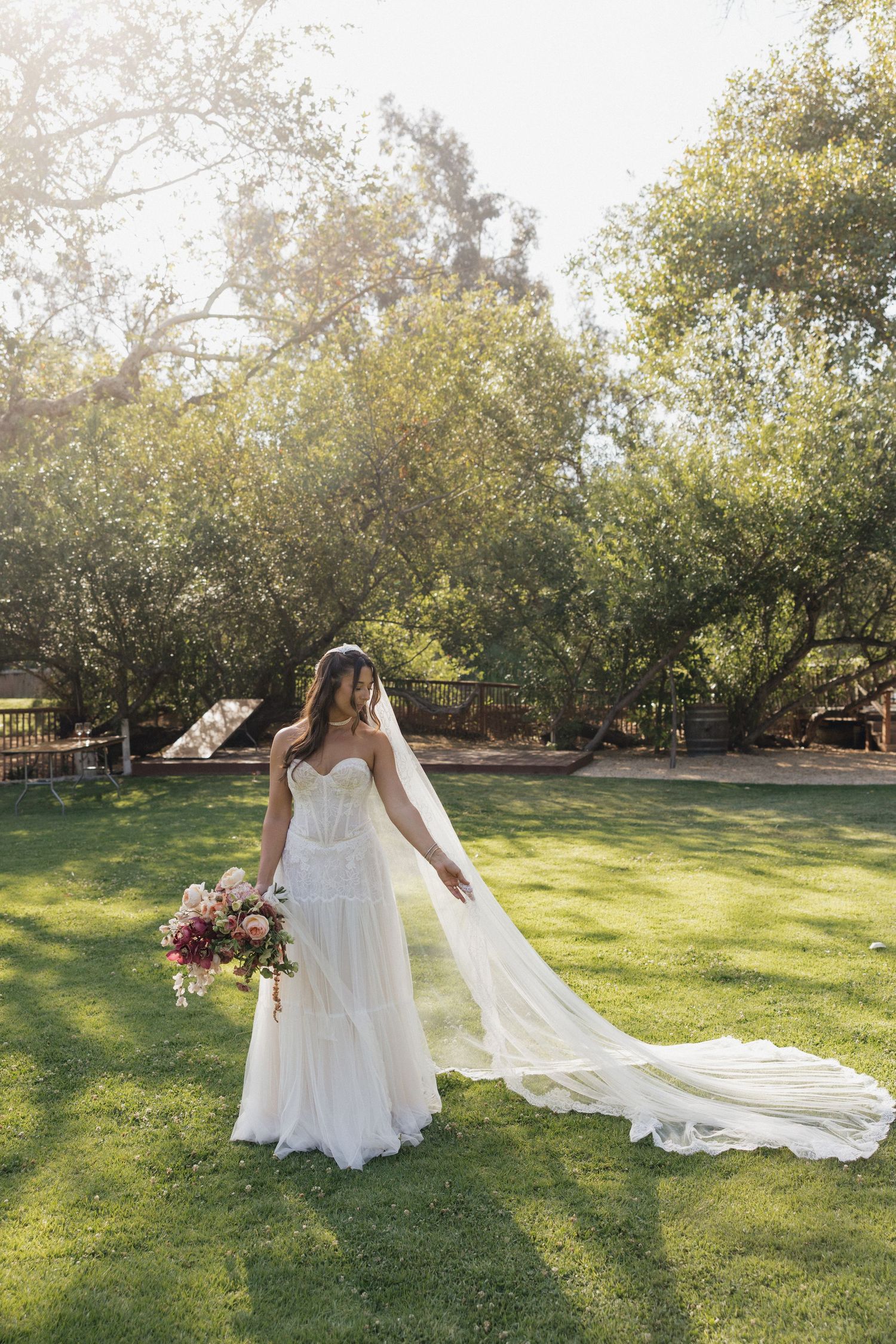 Elegant bride in lace gown with cathedral veil holds bouquet on sunlit green lawn outdoors.