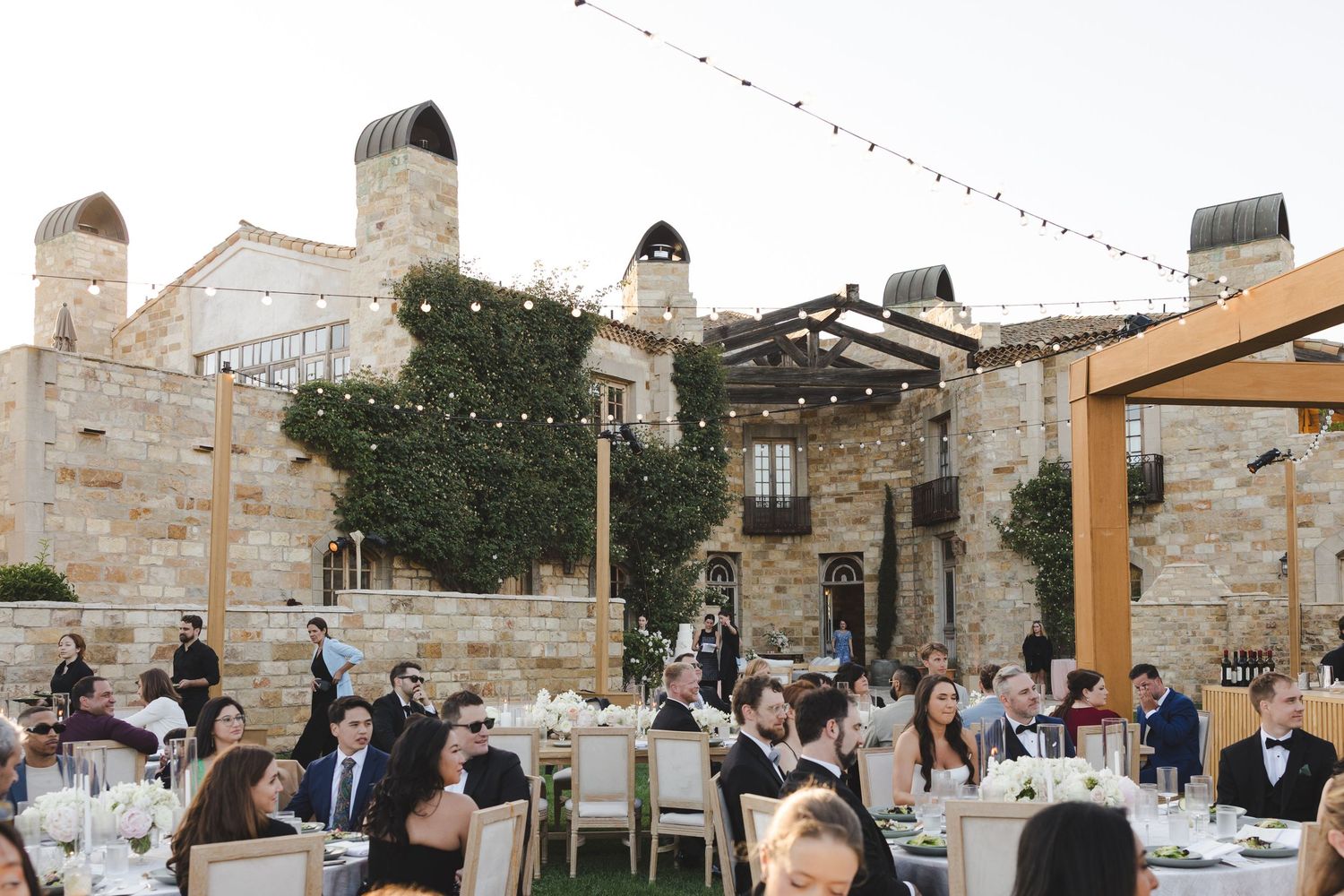 Wedding guests gather for an outdoor reception at a rustic stone venue with string lights overhead.