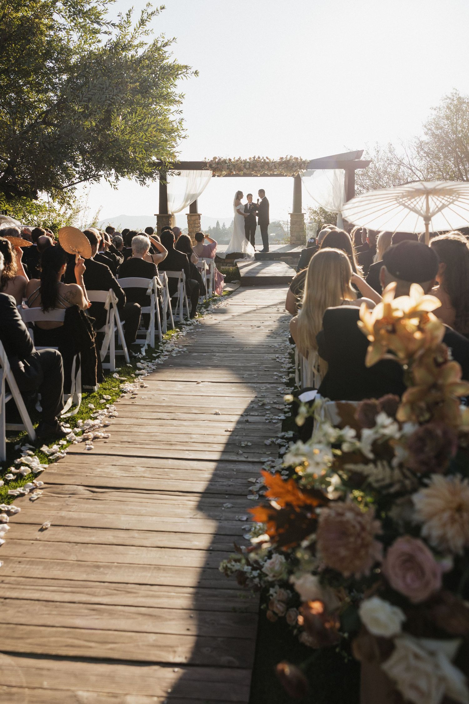 Outdoor wedding ceremony on a wooden deck with floral arrangements and guests seated under a rustic arbor.