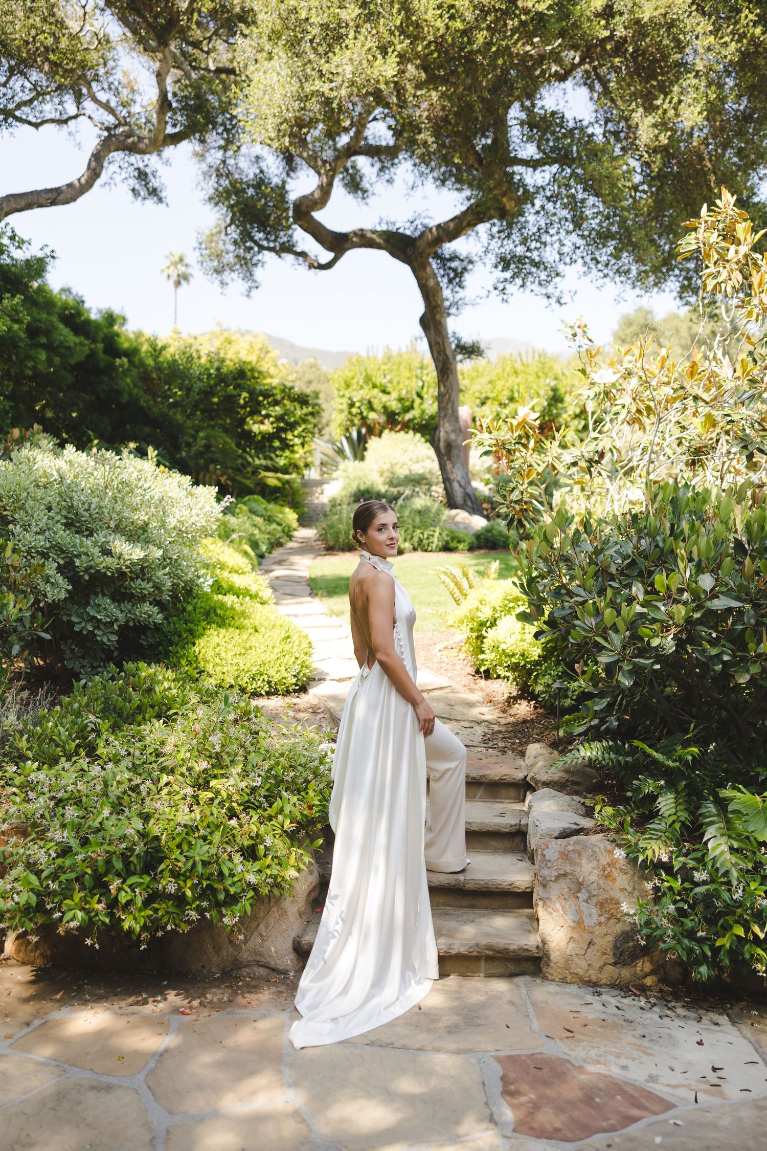 Figure in flowing white dress twirls gracefully among garden pathways and lush greenery on a sunny afternoon.