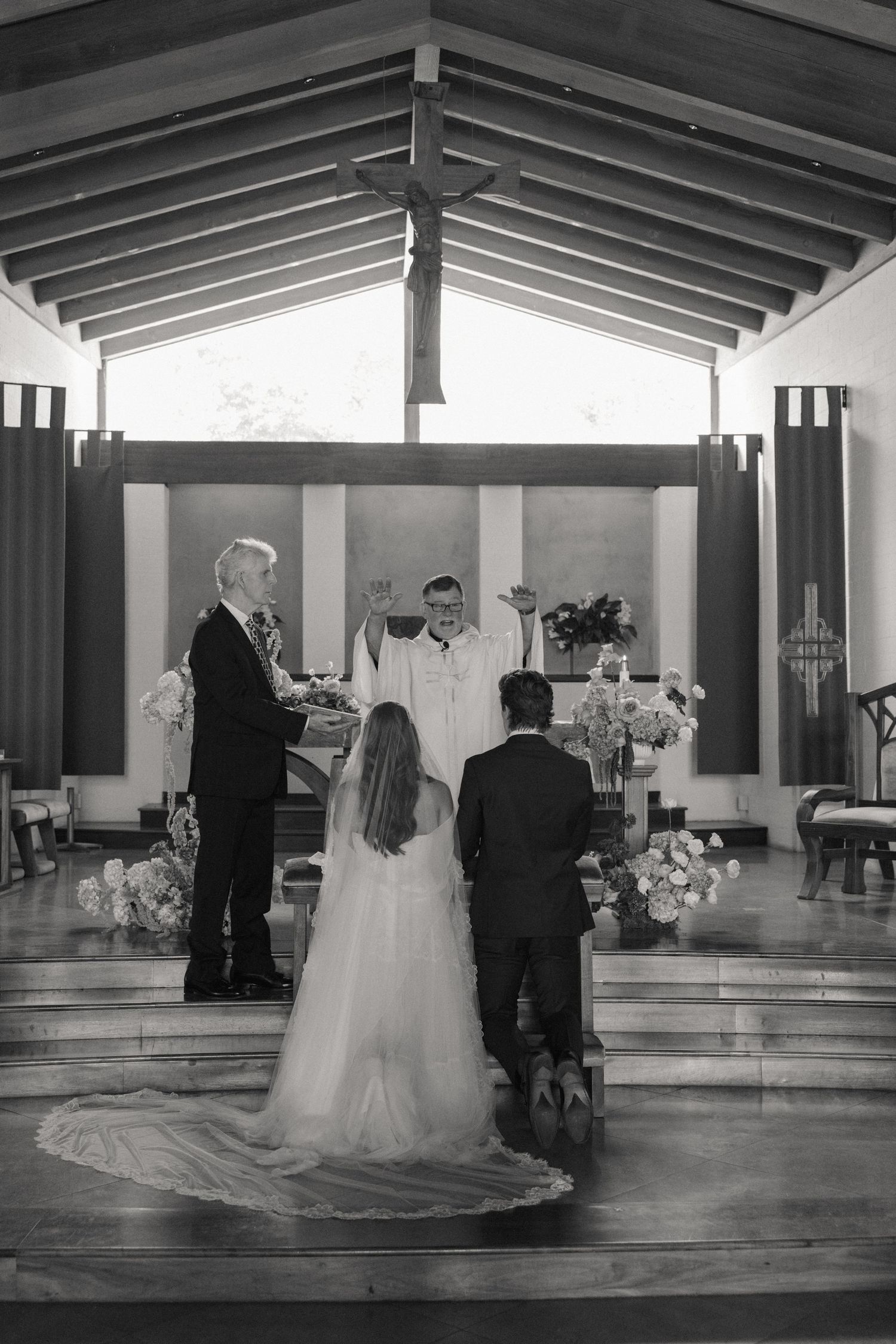 Black and white photo of bride and groom at church altar with priest raising hands during wedding ceremony.
