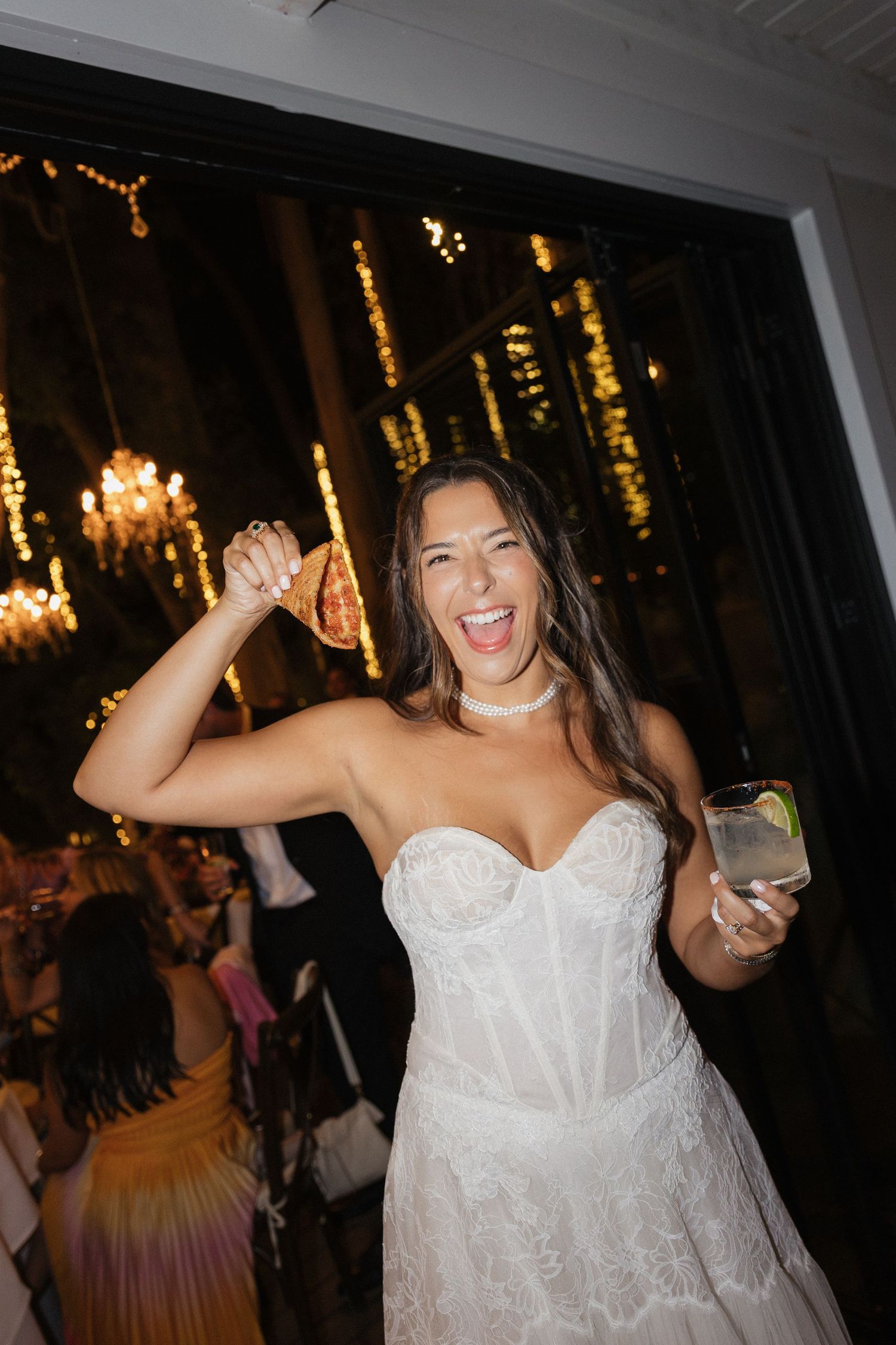 Excited bride in strapless lace gown holding pizza slice and cocktail glass at wedding reception.