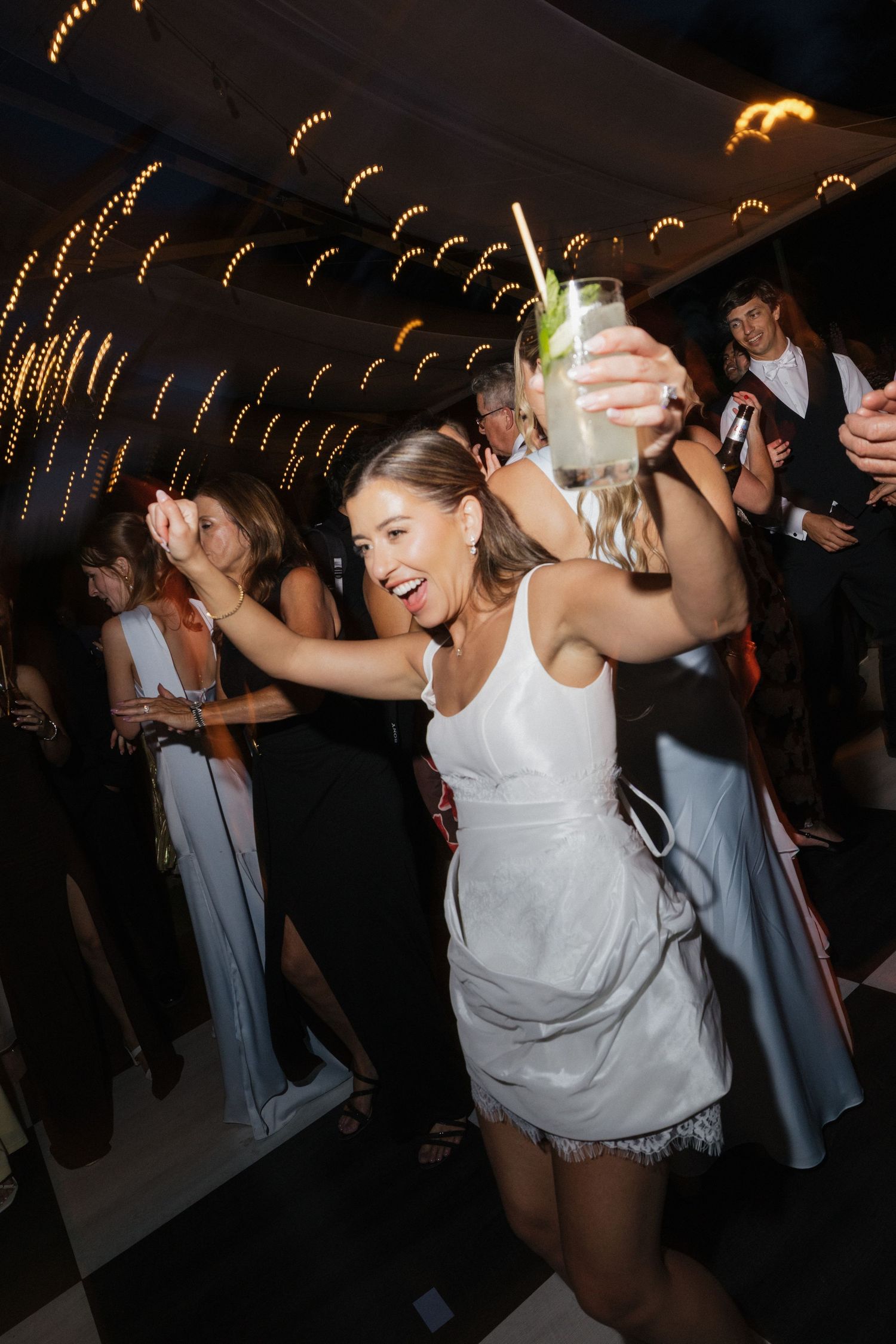 Woman in white dress dancing joyfully at a party, holding a cocktail glass, with guests in the background.