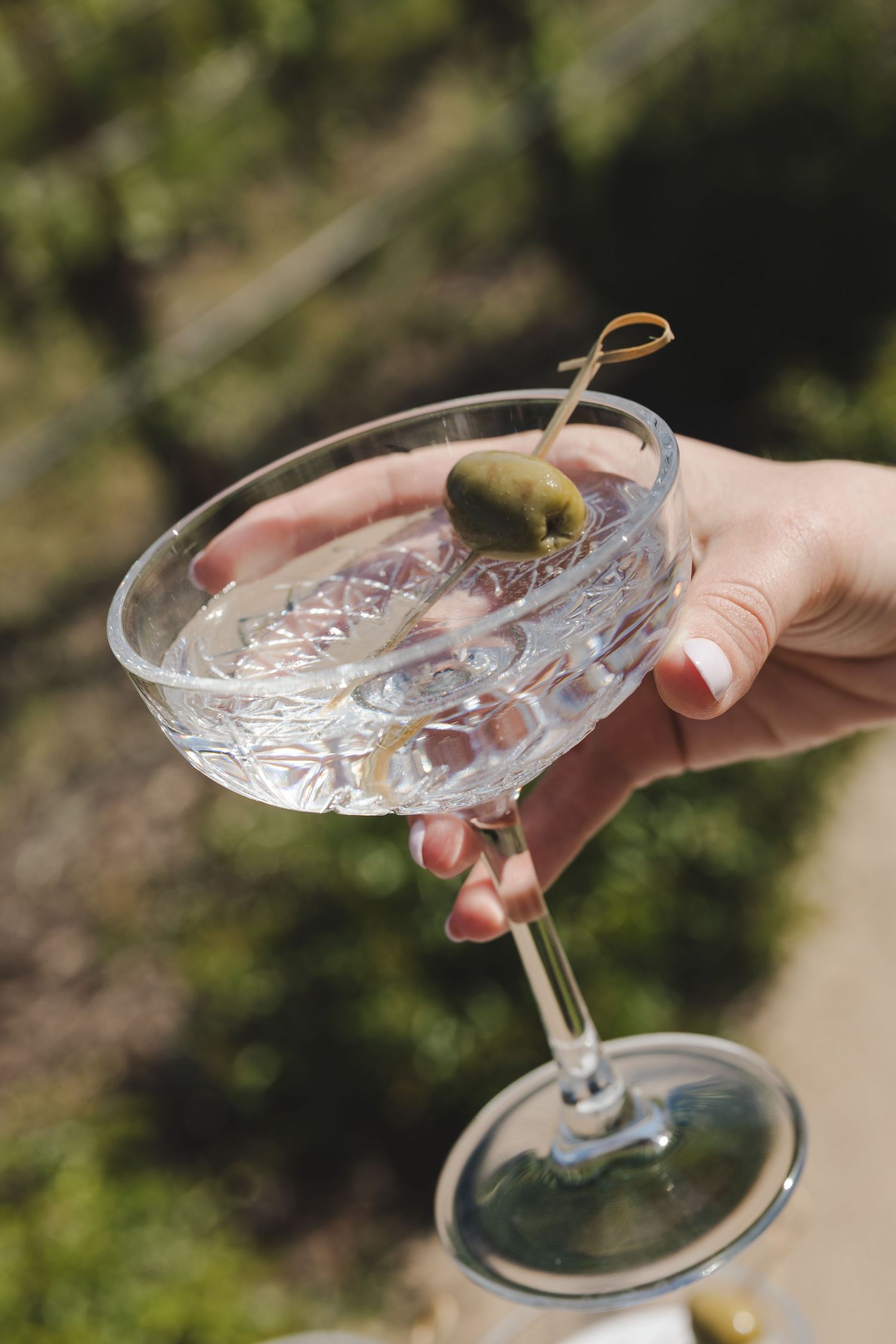 A hand holds an elegant crystal martini glass with an olive garnish against a blurred outdoor background.