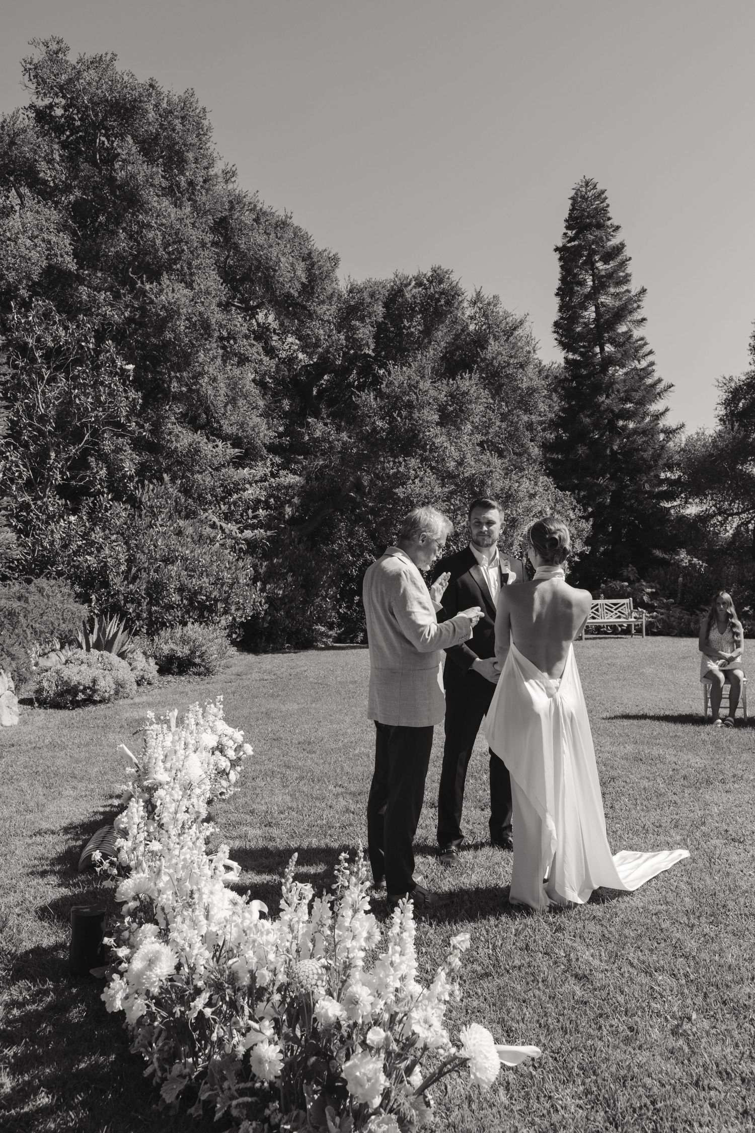 Black and white outdoor wedding ceremony takes place on a grassy hillside with trees in the background.