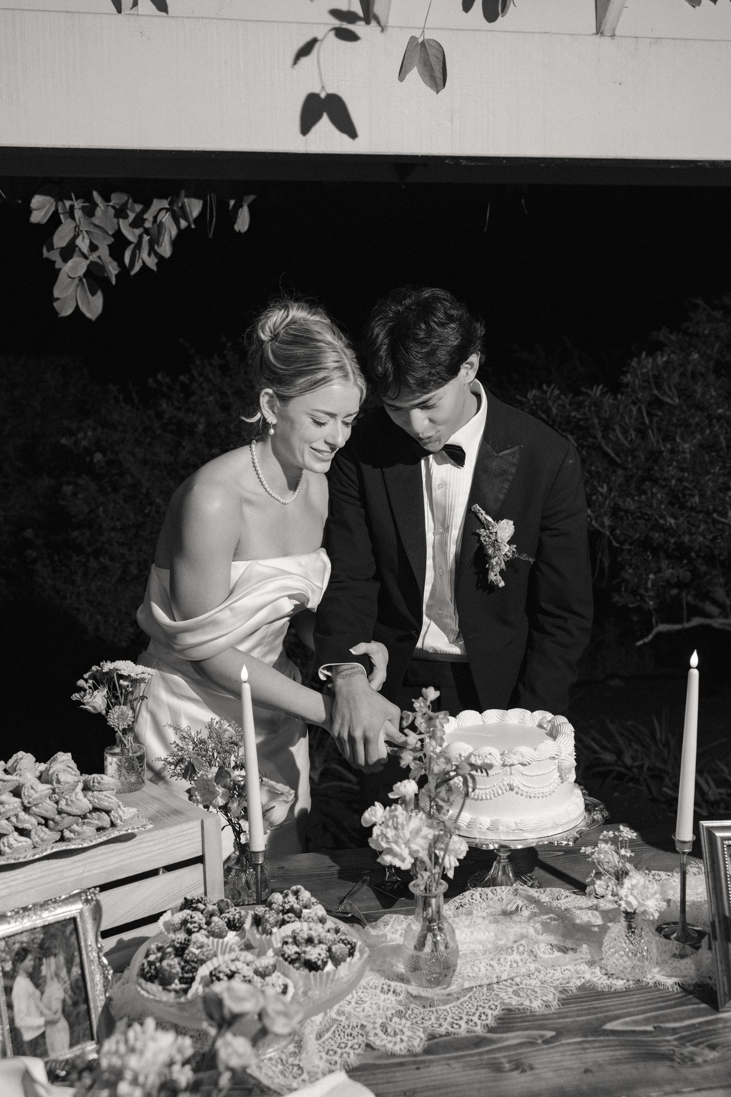 A couple cuts their elegant white wedding cake together at a reception decorated with floral arrangements and candles.