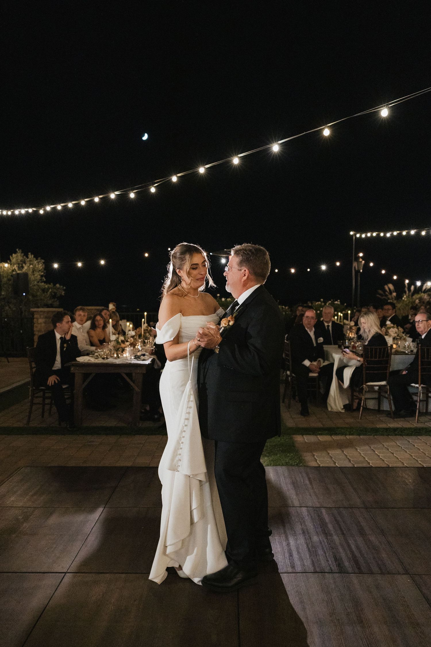 A couple shares their first dance at an outdoor evening wedding reception under romantic string lights.