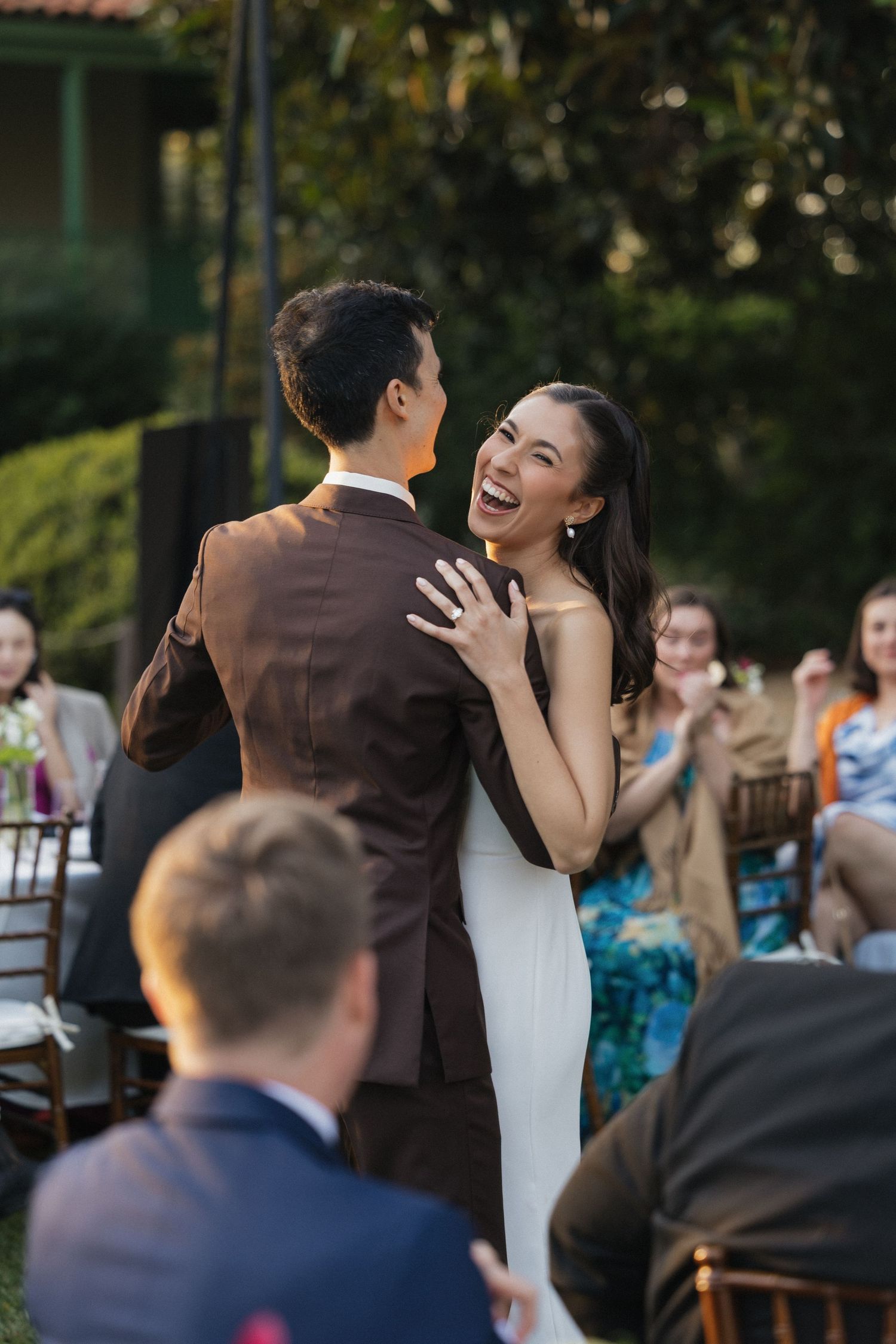 Joyful bride in white gown laughs while sharing first dance with groom in brown suit at outdoor wedding reception.