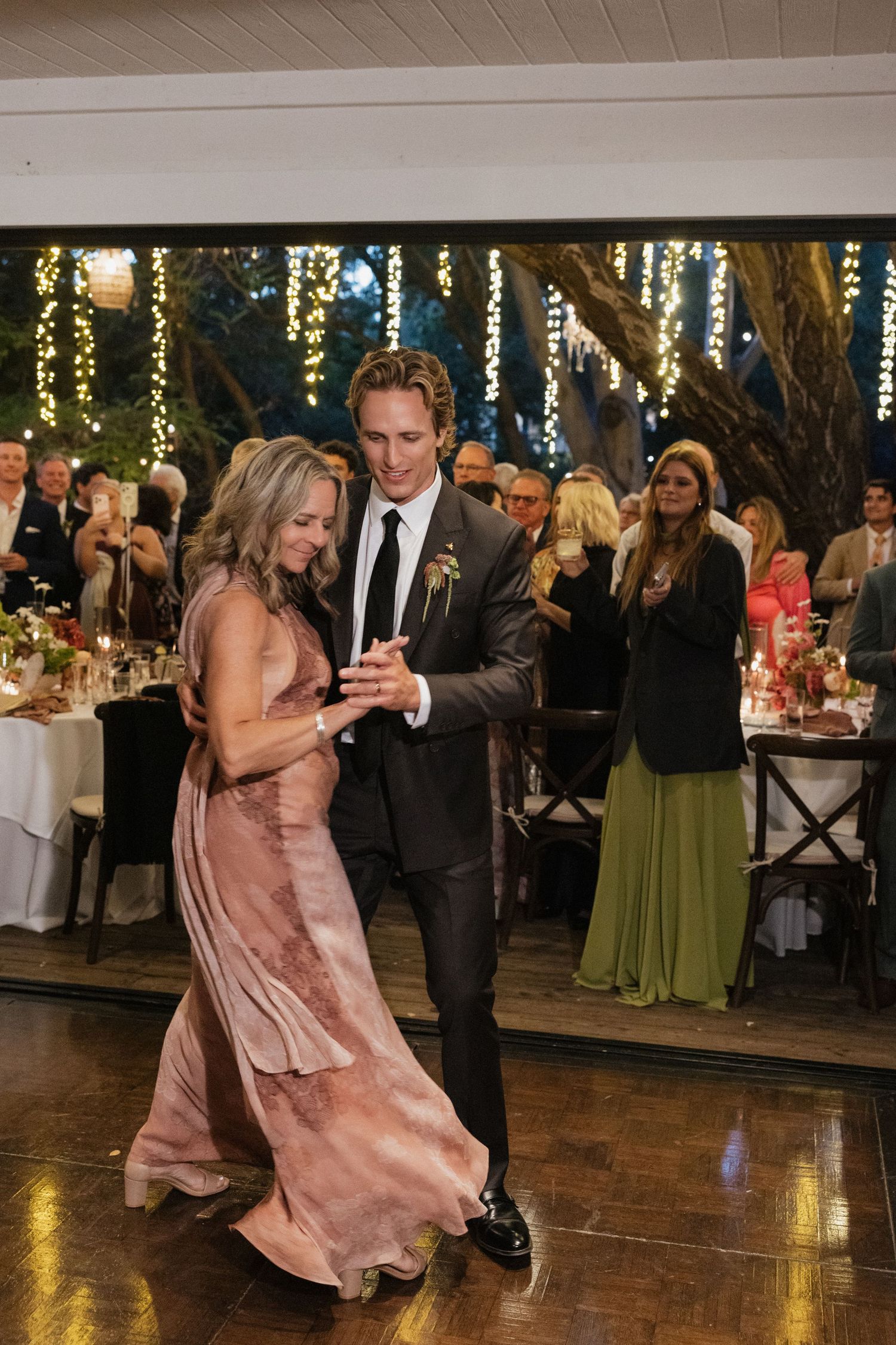 Groom dances with mother in a dusty rose gown at a romantic wedding reception with fairy lights.