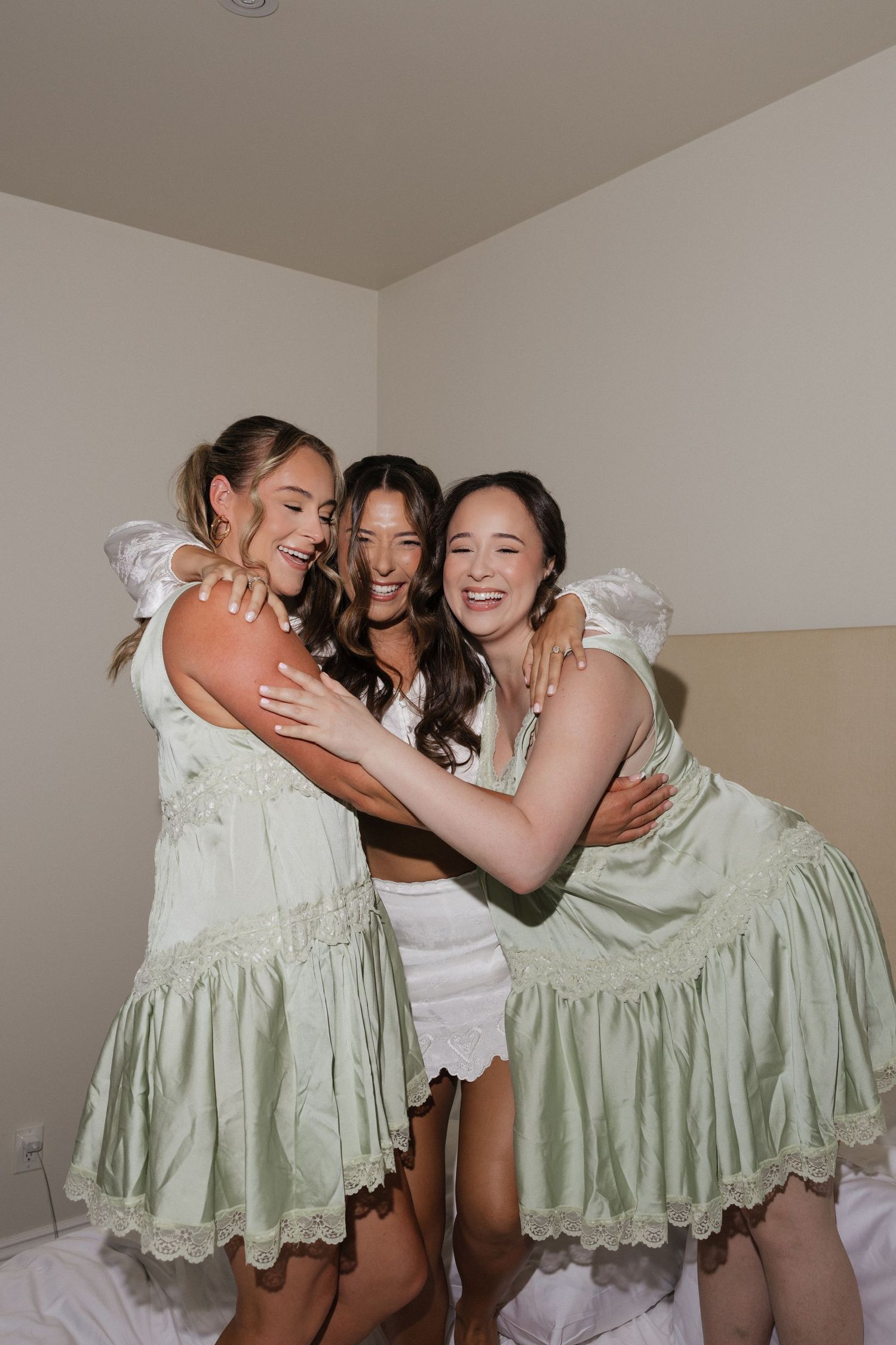 Three women in sage green dresses embrace and laugh together in a bright indoor setting.