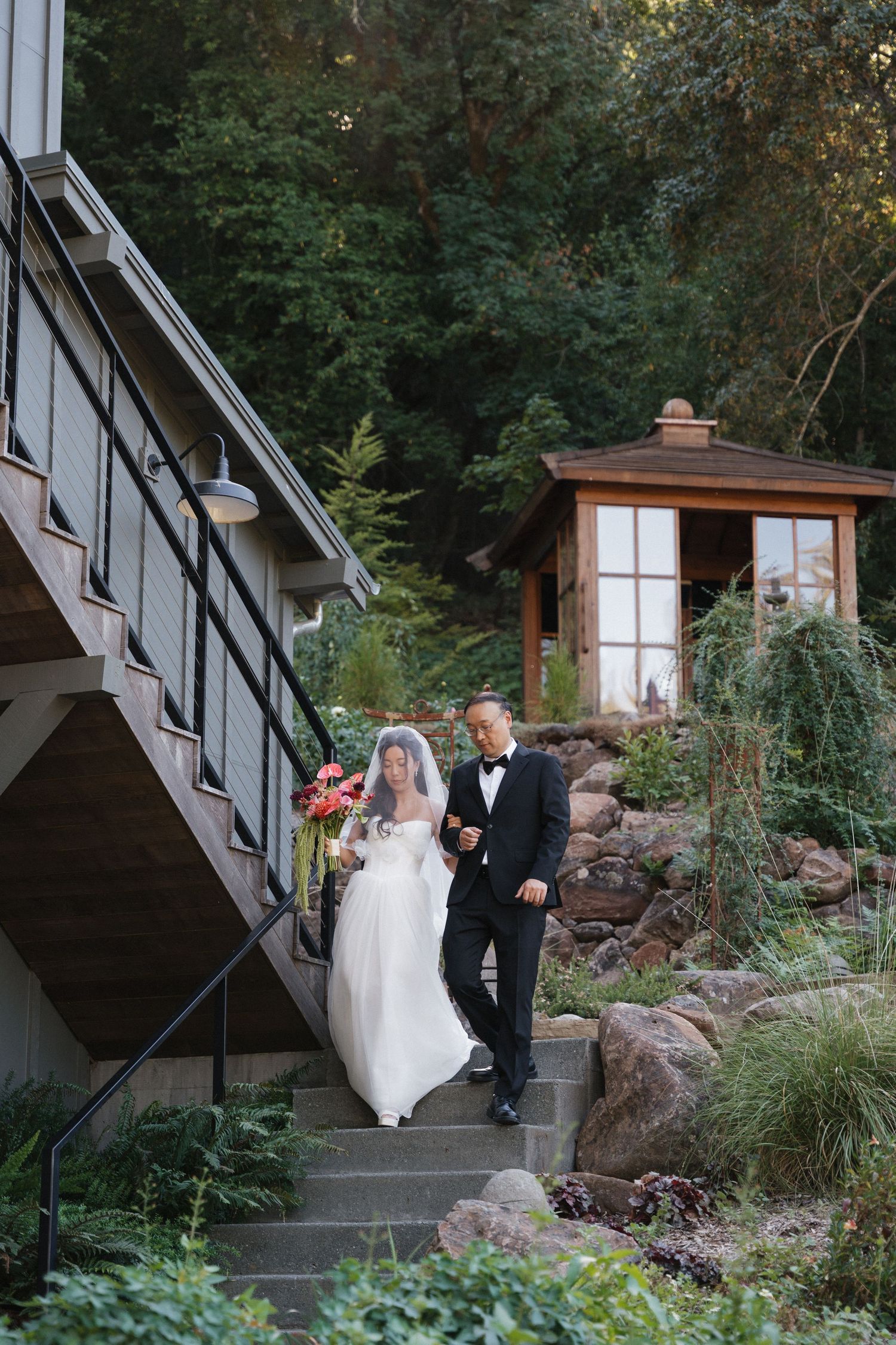 A couple in wedding attire walks together on stone steps surrounded by lush greenery.
