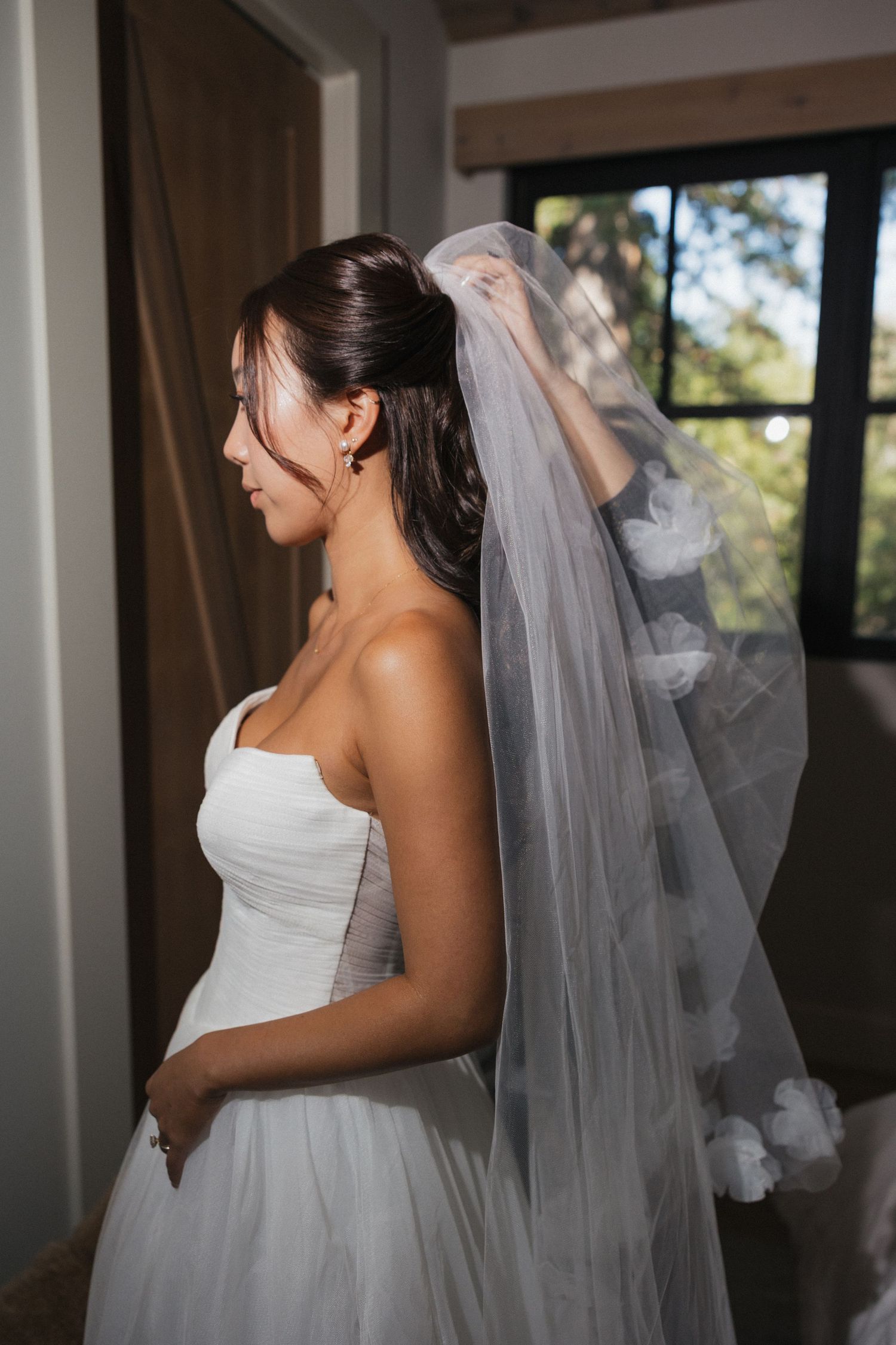 A bride in a white wedding gown and veil getting ready in a sunlit room.