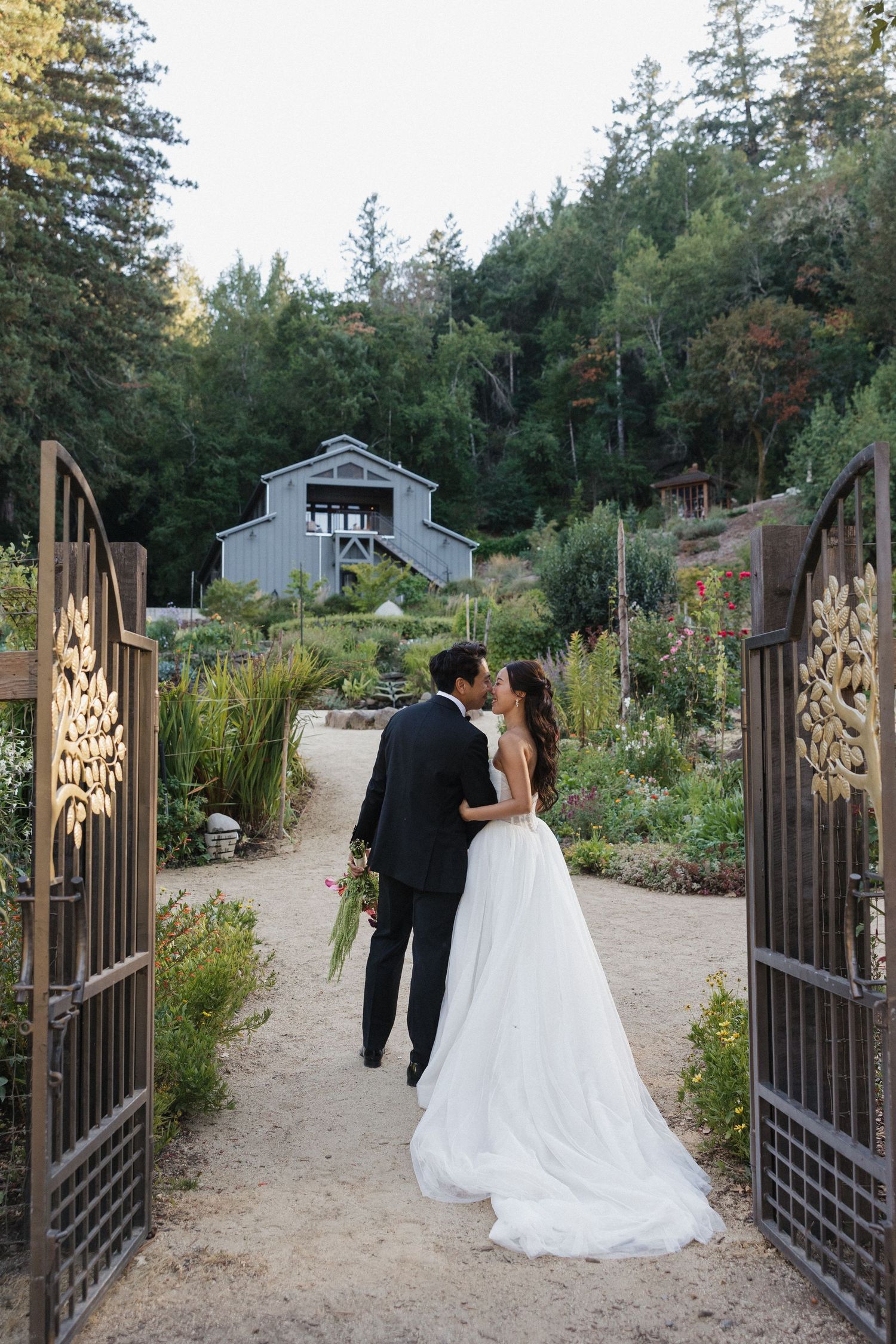 Couple walking through ornate garden gates on path surrounded by trees.