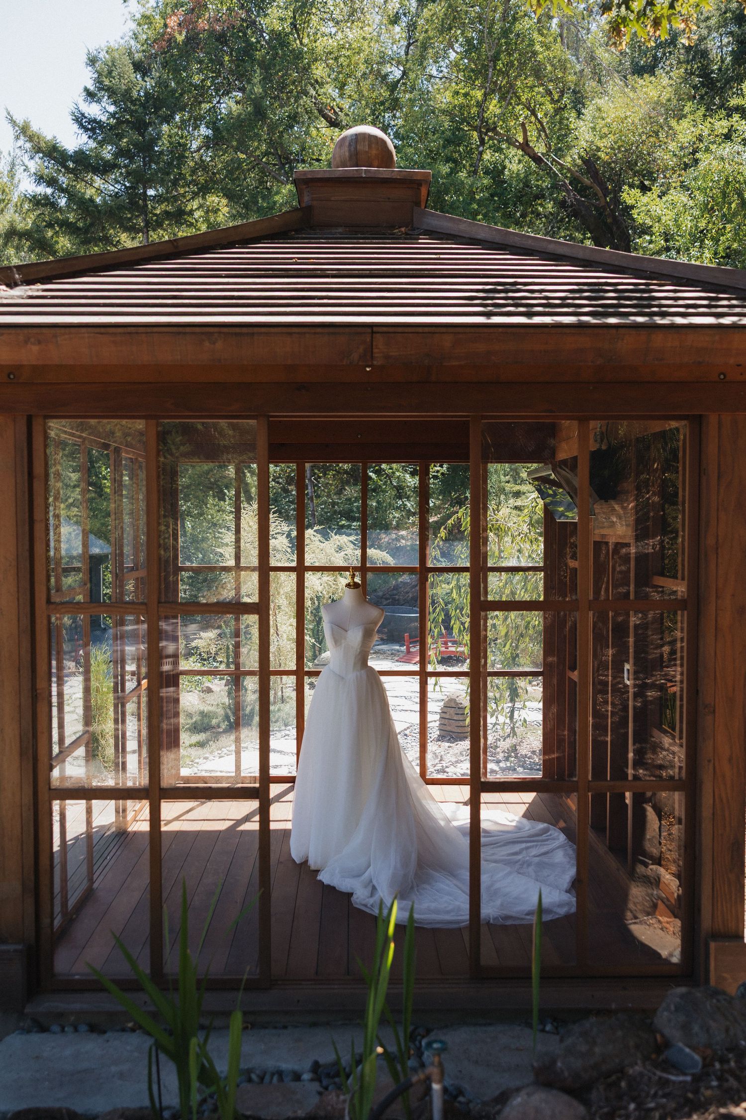 A series of wedding dresses displayed in a rustic wooden gazebo with glass windows surrounded by trees and natural light.