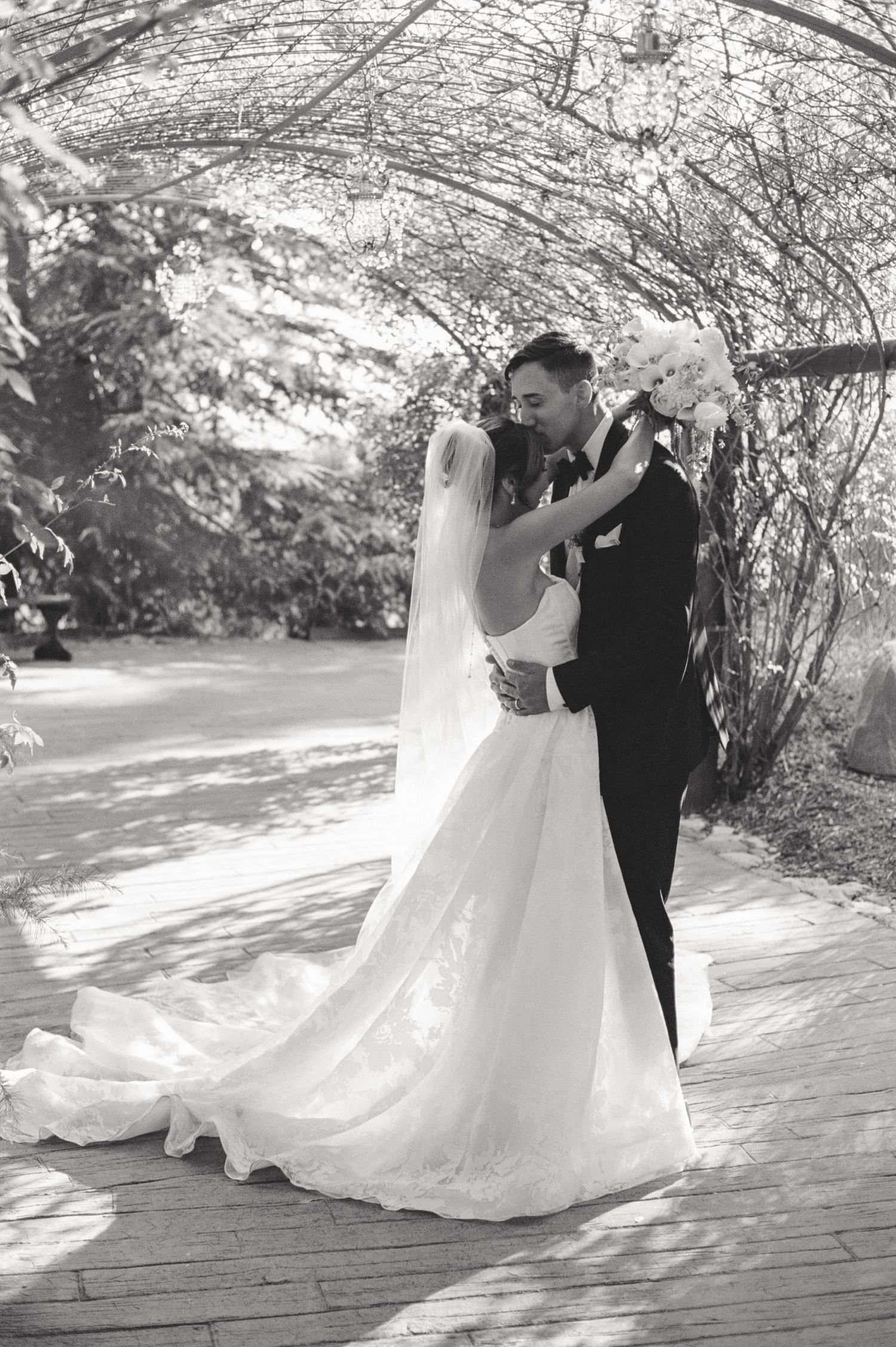 Romantic black and white wedding photo of newlyweds embracing under flowering tree branches on wooden deck.