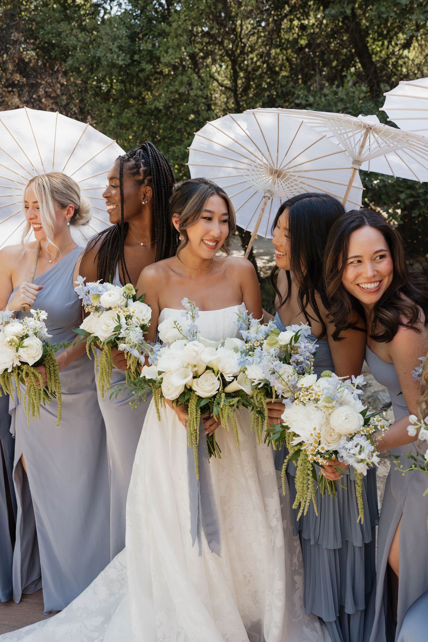 Bridesmaids in grey dresses holding white parasols and bouquets pose together at an outdoor wedding.
