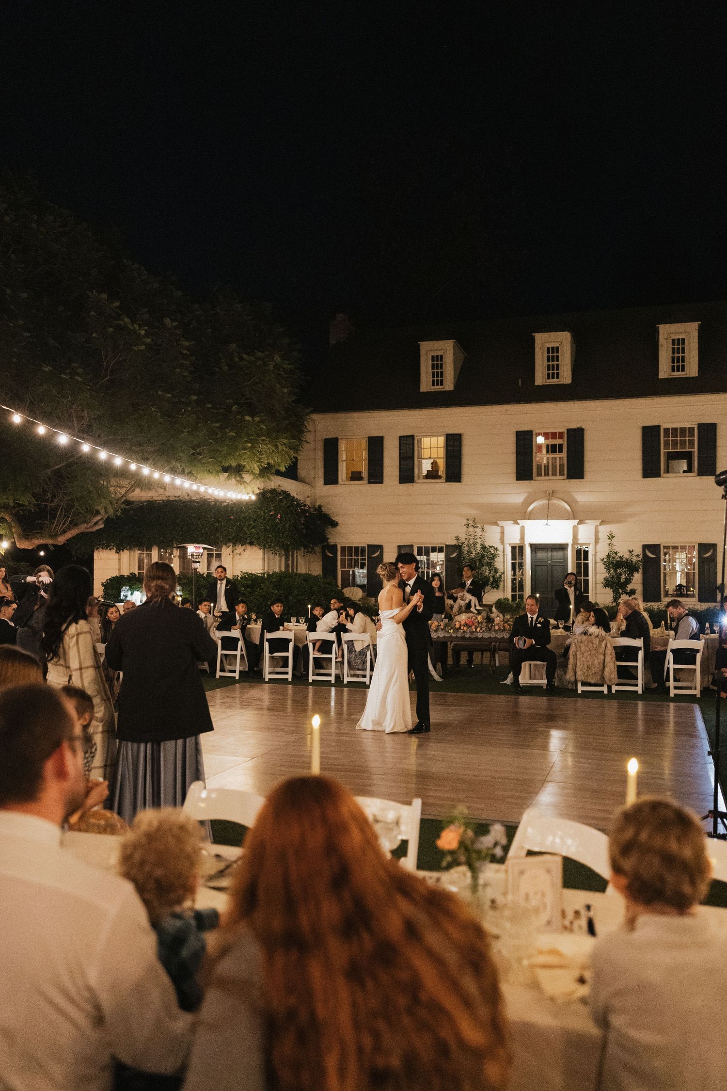 Guests watch a couple's first dance at an evening wedding reception under string lights and white decor.