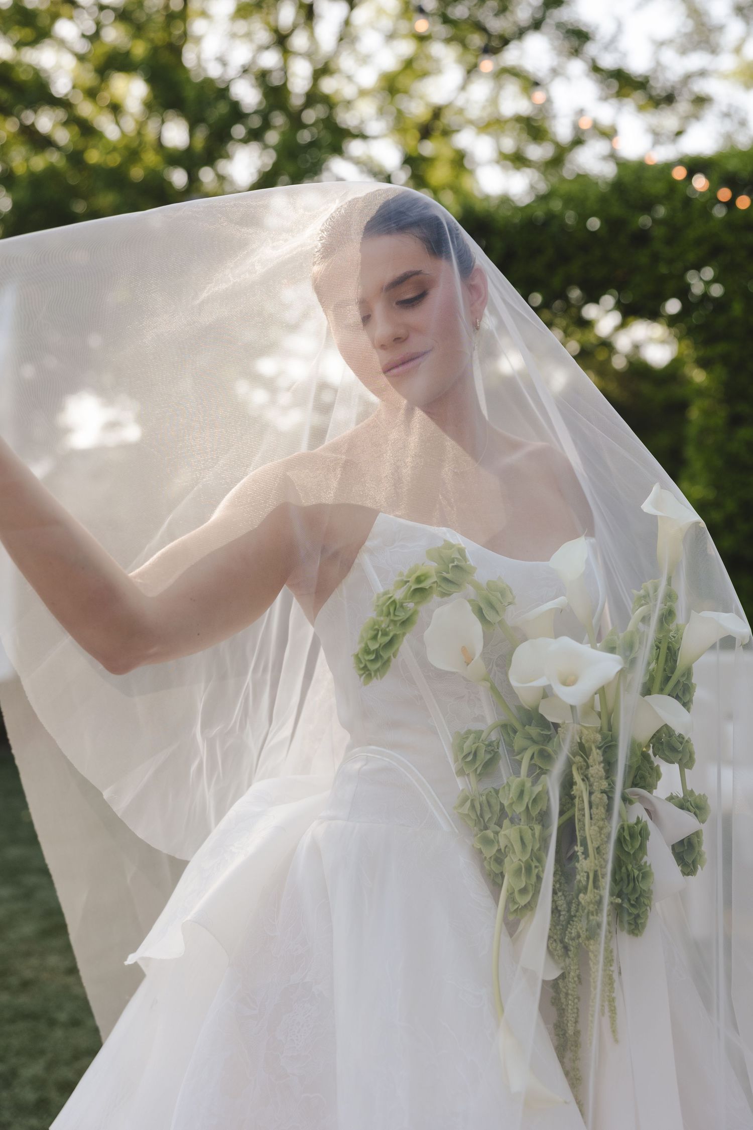 A bride in white wedding dress and veil holds a bouquet of white flowers in a sunny outdoor garden setting.