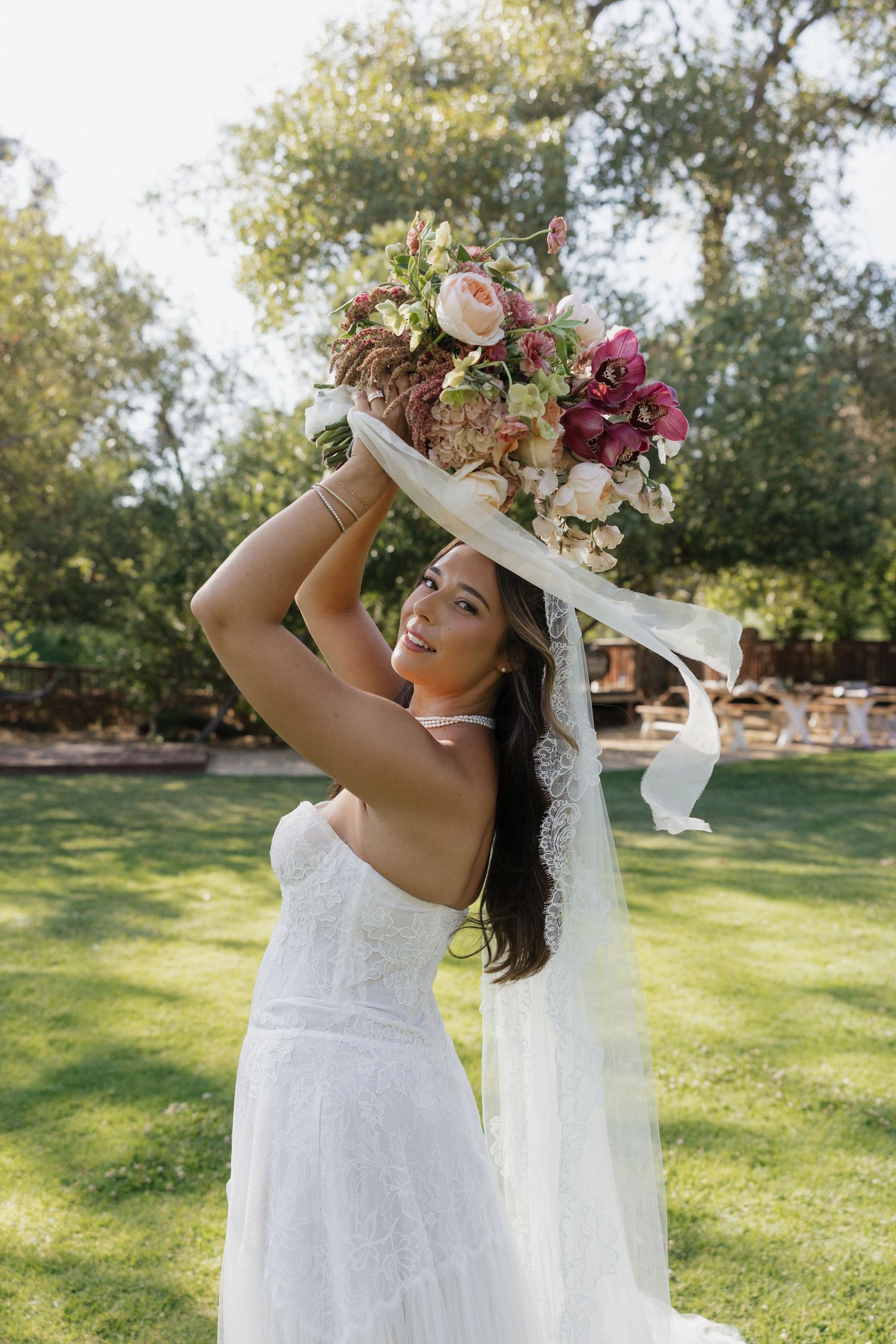 Bride in white lace gown holds colorful floral bouquet overhead while posing outdoors on green lawn.