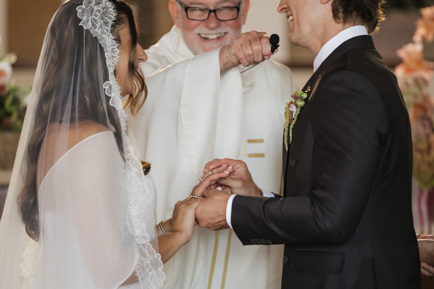 Bride and groom exchange rings during wedding ceremony with officiant smiling in background.