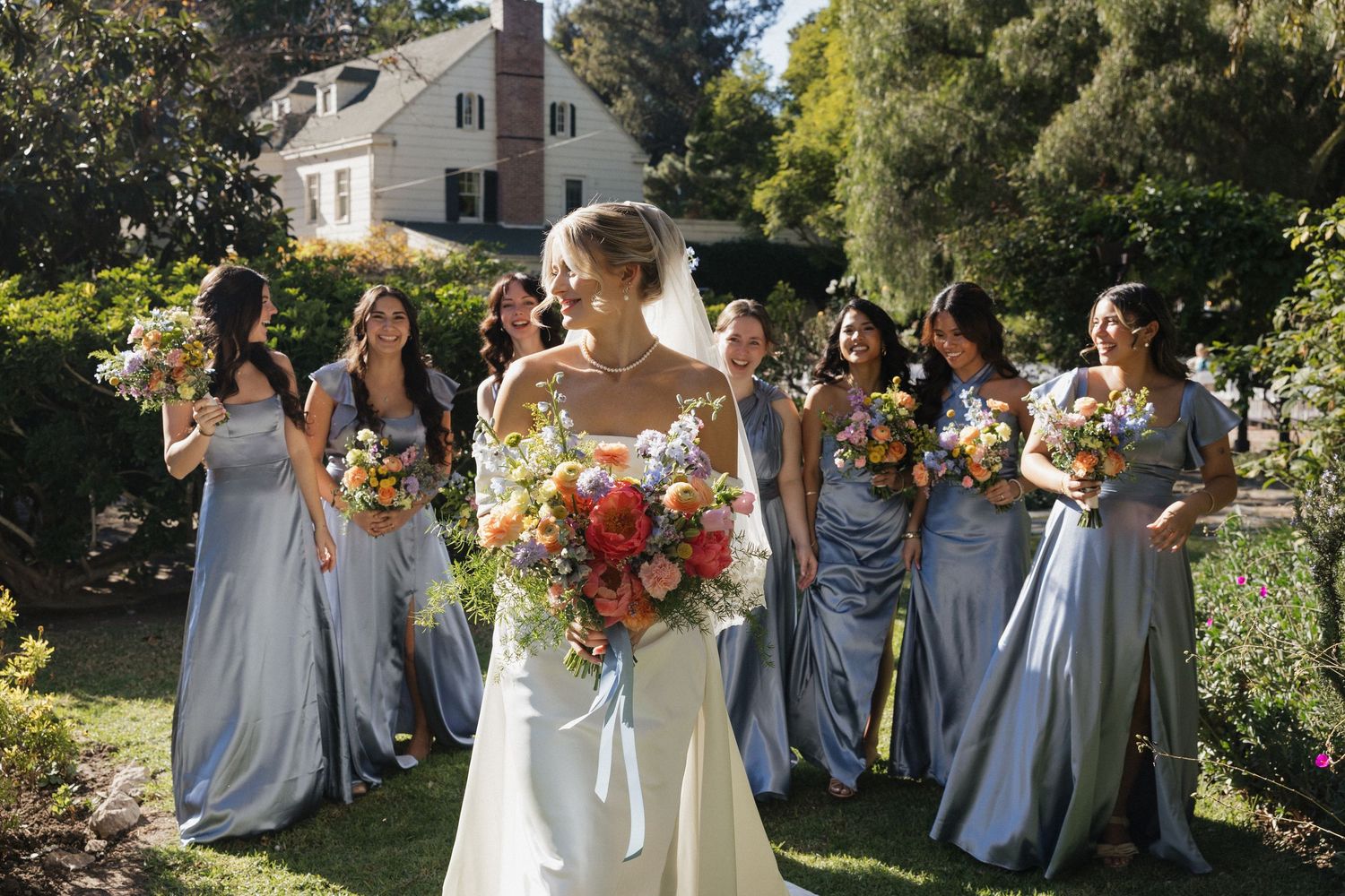 Wedding party in light blue bridesmaid dresses poses together in a sunny garden setting with vibrant flowers.
