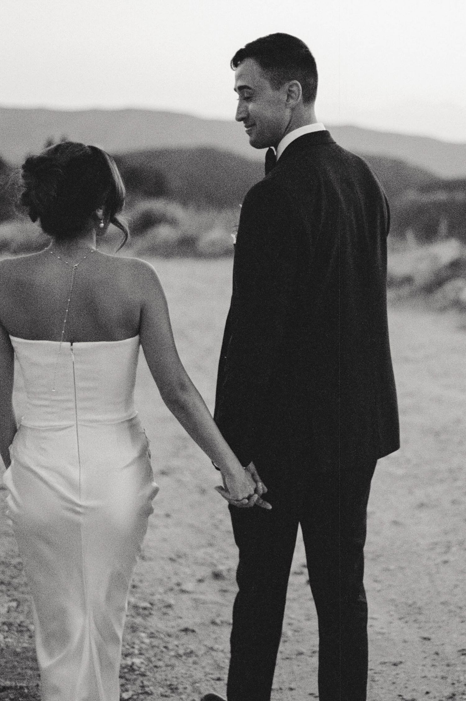 Black and white portrait of couple in wedding attire holding hands against mountainous backdrop.
