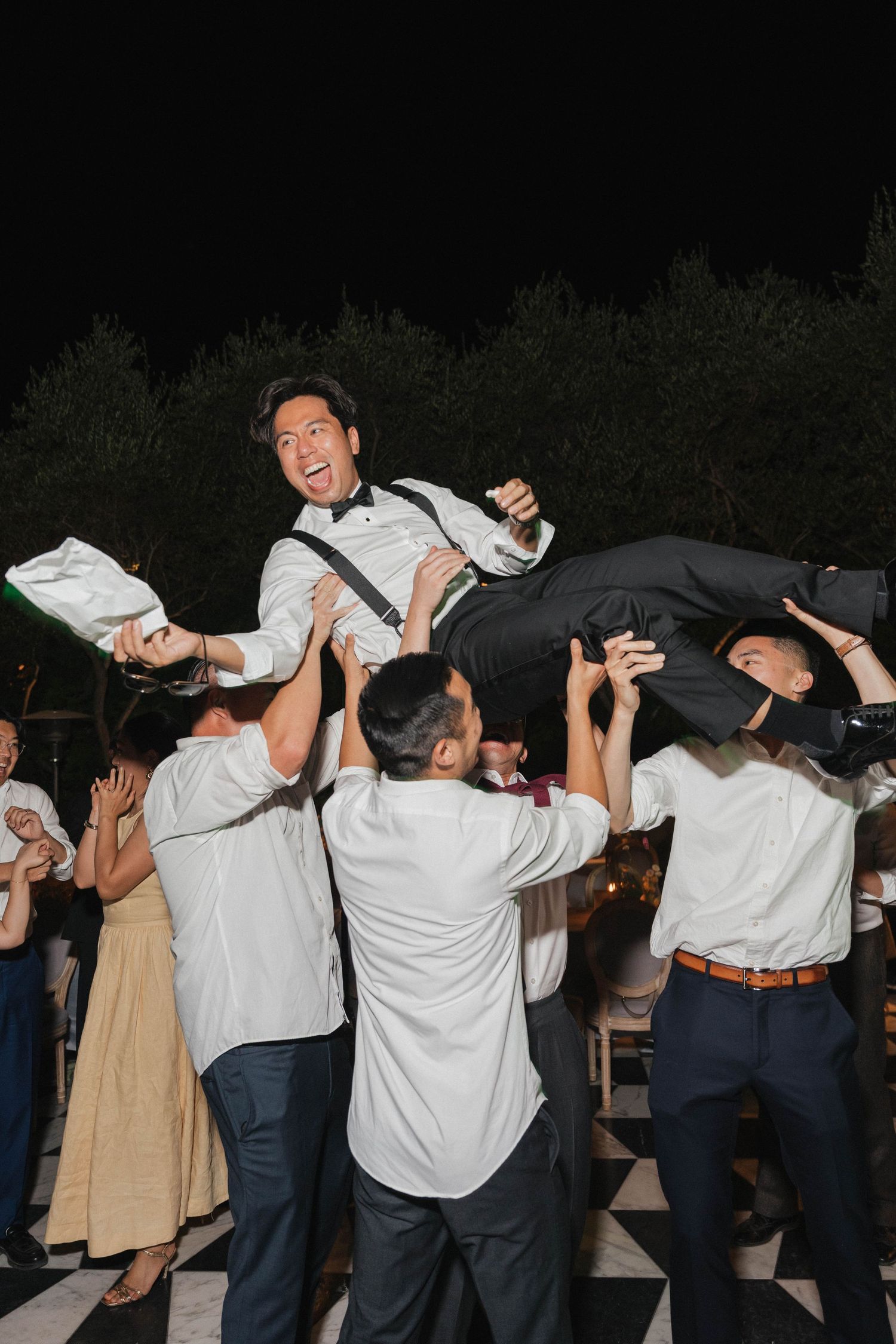 Group performs crowd surfing at nighttime party wearing white shirts and casual attire.