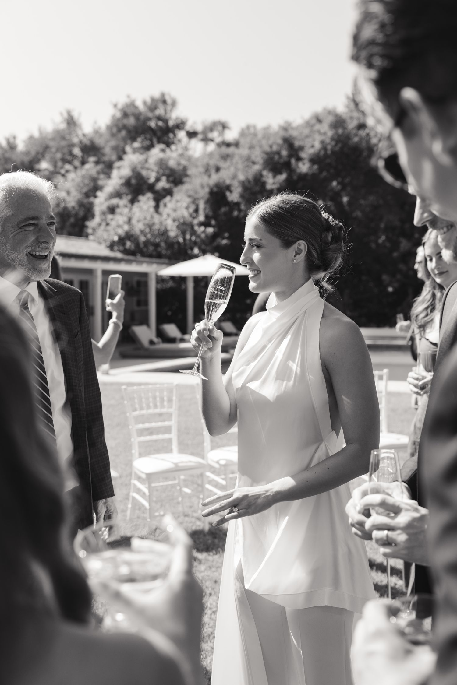 Wedding guests dressed in formal attire gather outdoors around elegant white chairs during a daytime celebration.
