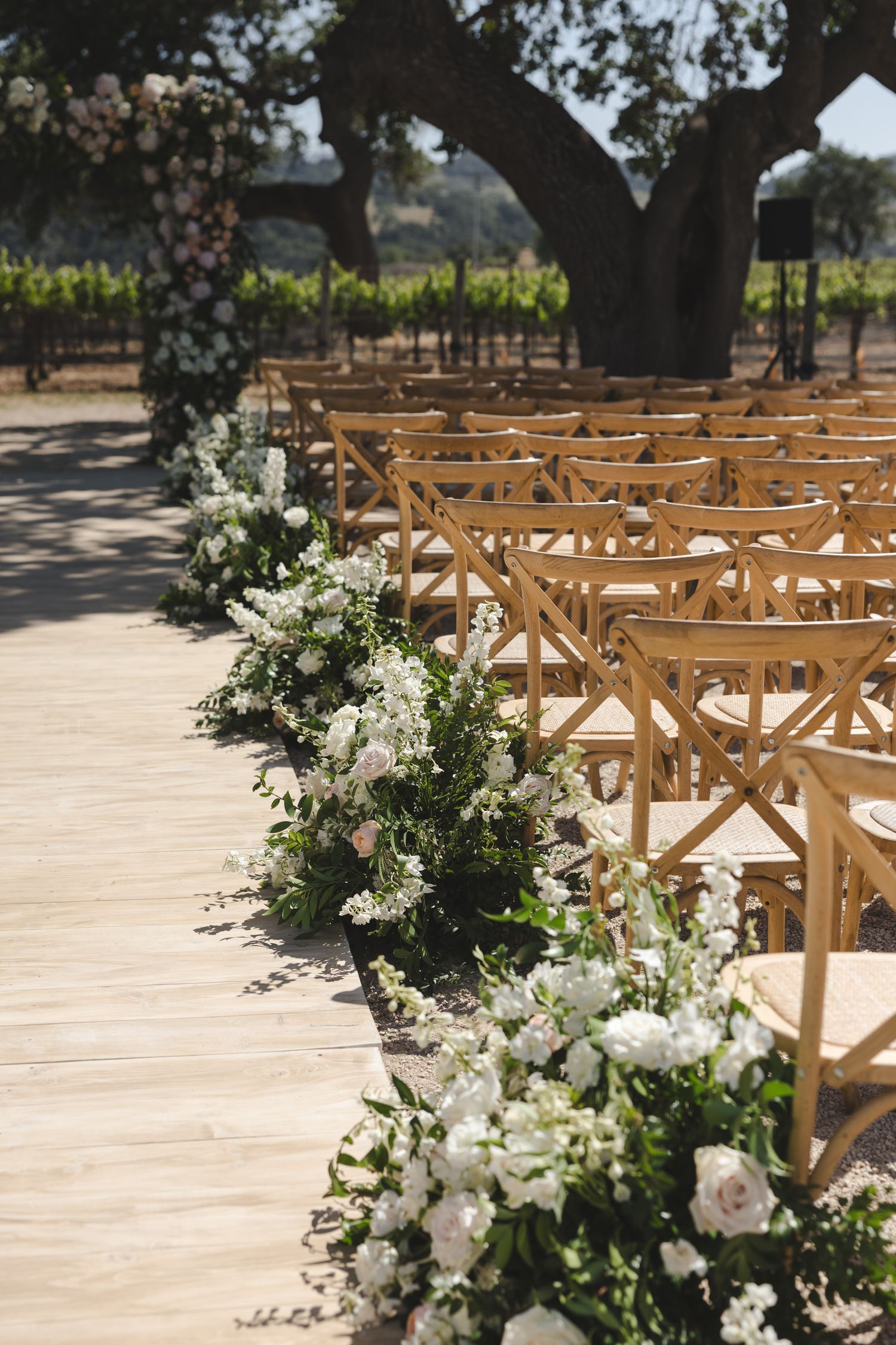 Wooden cross-back chairs arranged for an outdoor wedding ceremony with white floral decorations along the aisle.