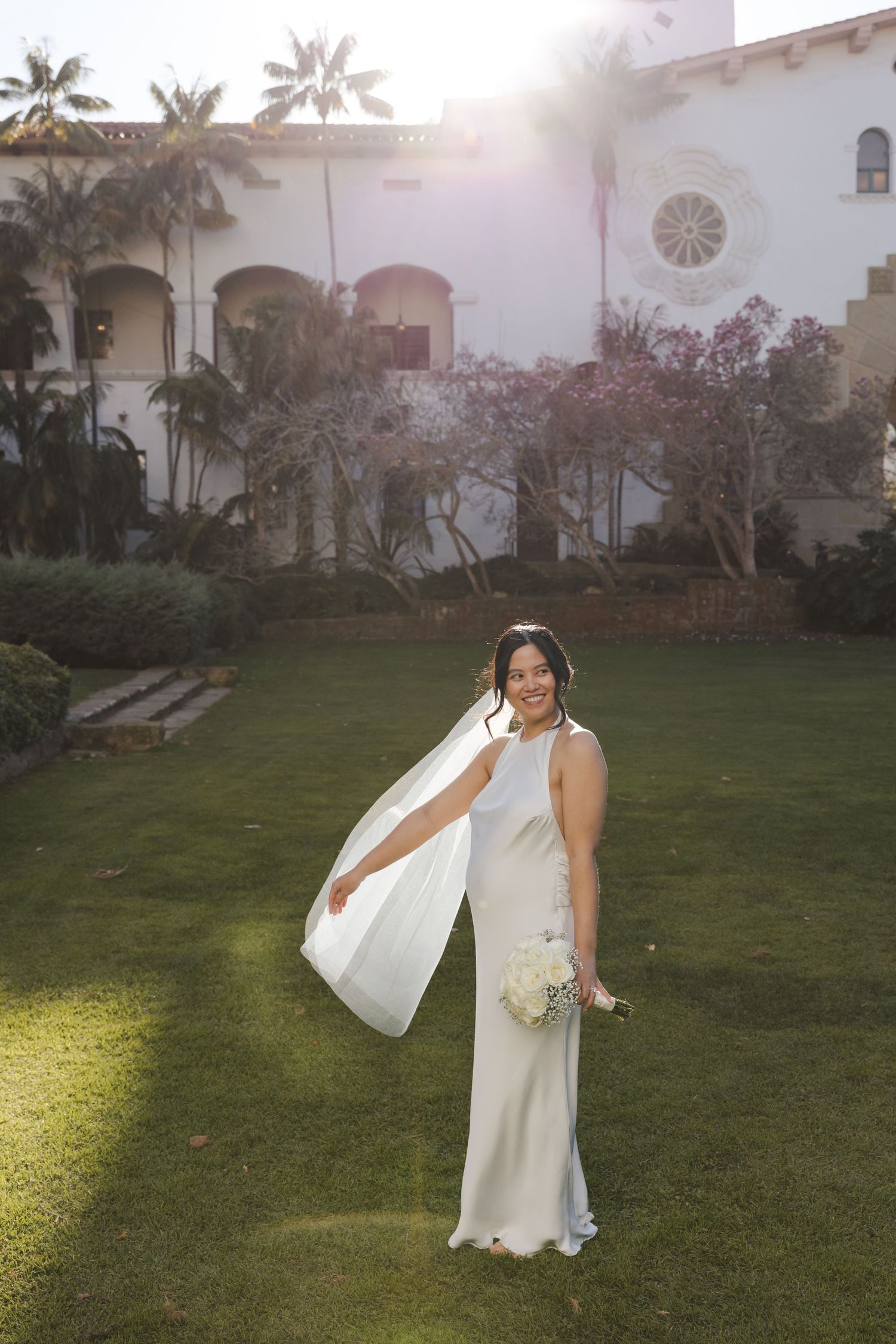 A bride in a sleek white dress poses on a lawn with a Spanish-style building and palm trees in the background.