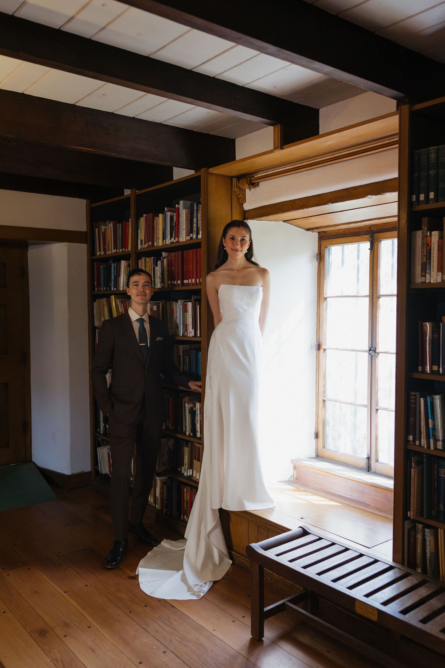 Bride in strapless white gown stands by window in wood-paneled library as groom in dark suit looks on.