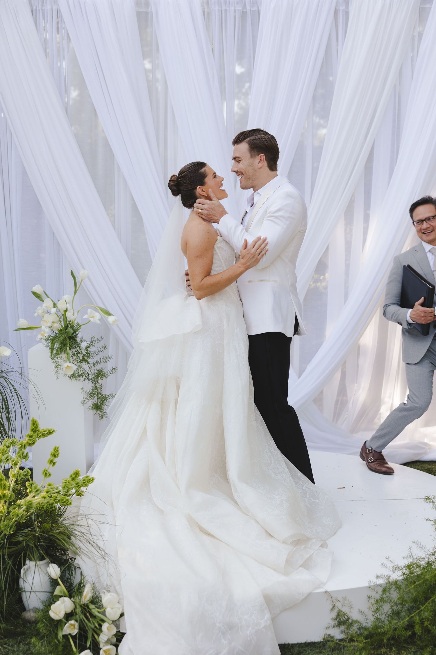 A bride and groom embrace during their wedding ceremony surrounded by white drapes and greenery decorations.