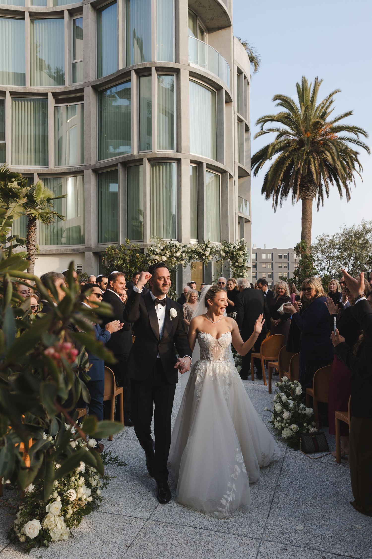 Elegant outdoor wedding ceremony taking place at a modern building with curved glass windows and palm trees at sunset.