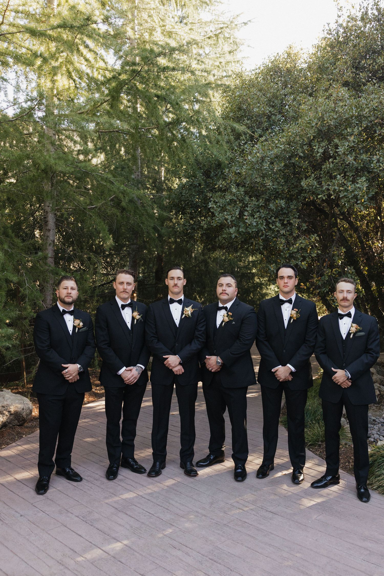 Groomsmen in black tuxedos and bow ties pose sitting together outdoors on stone steps during a wedding.