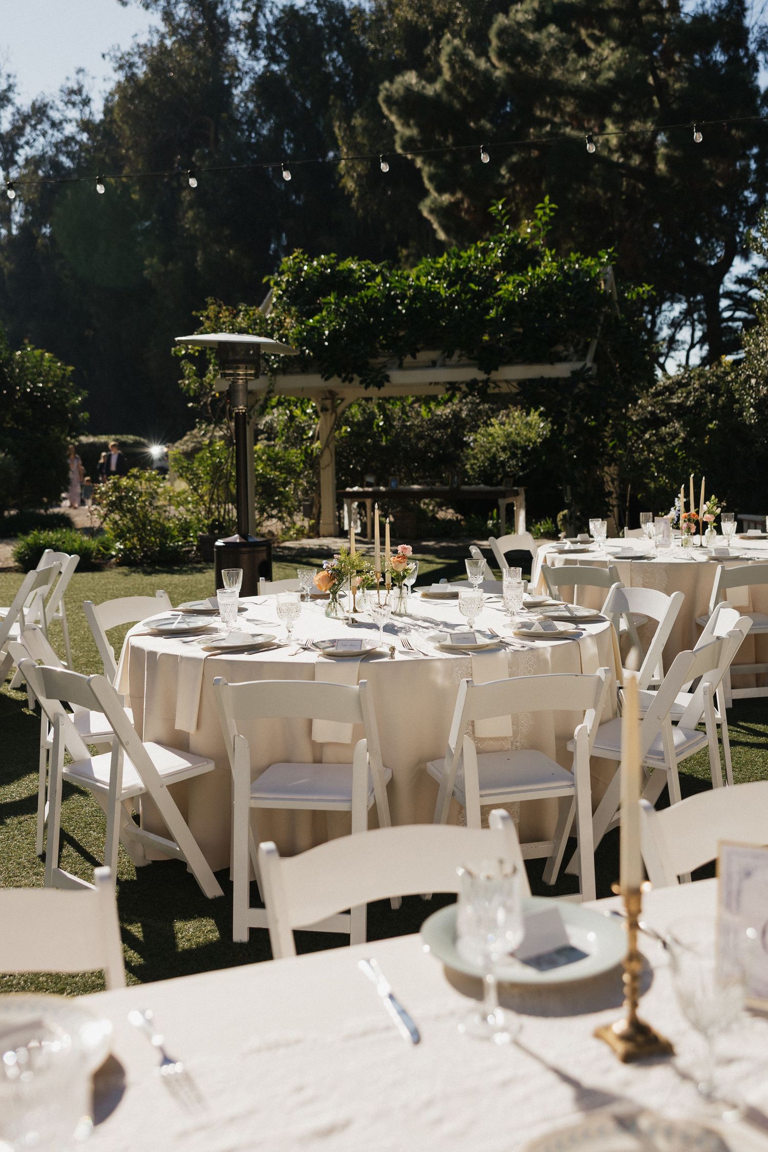 Elegant outdoor wedding reception setup with white tables, chairs and place settings in a garden setting.